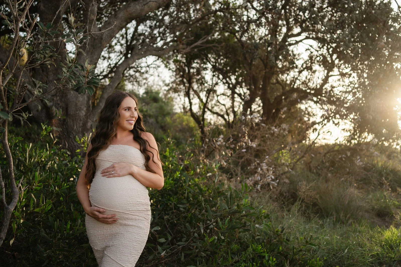 A pregnant woman in a beige strapless dress standing outdoors near trees and bushes, smiling and looking to her left during sunset.