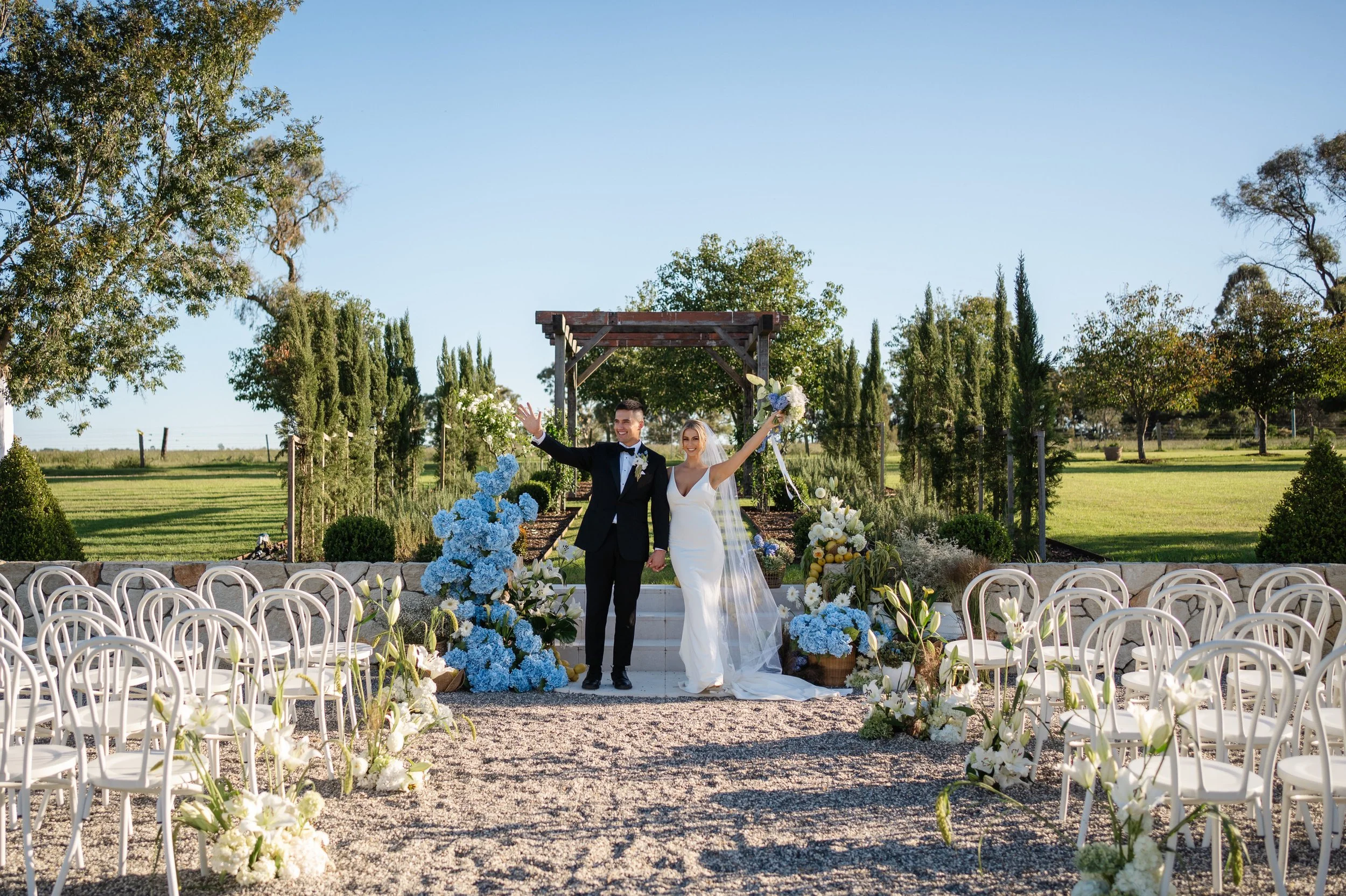 A bride and groom celebrating their wedding outdoors on a sunny day, holding hands with arms raised, surrounded by floral decorations and white chairs.