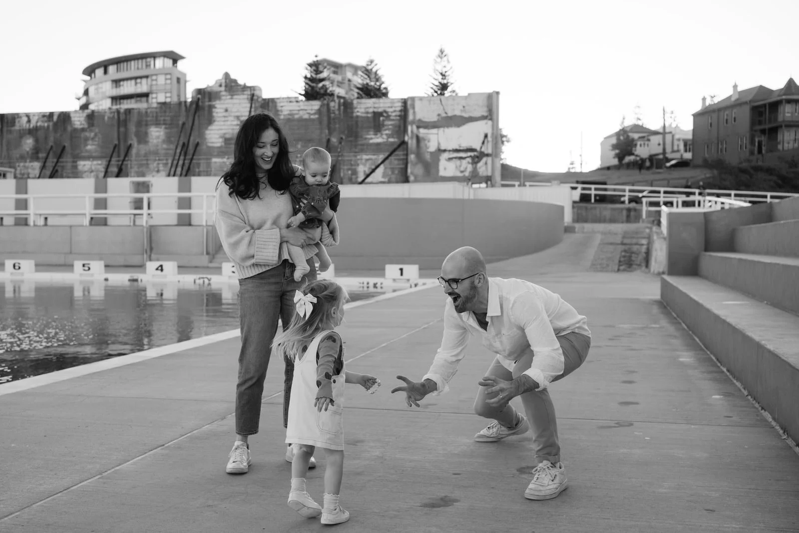 A happy family by a poolside, with two children and two adults, playing and smiling, in an outdoor setting near residential buildings.