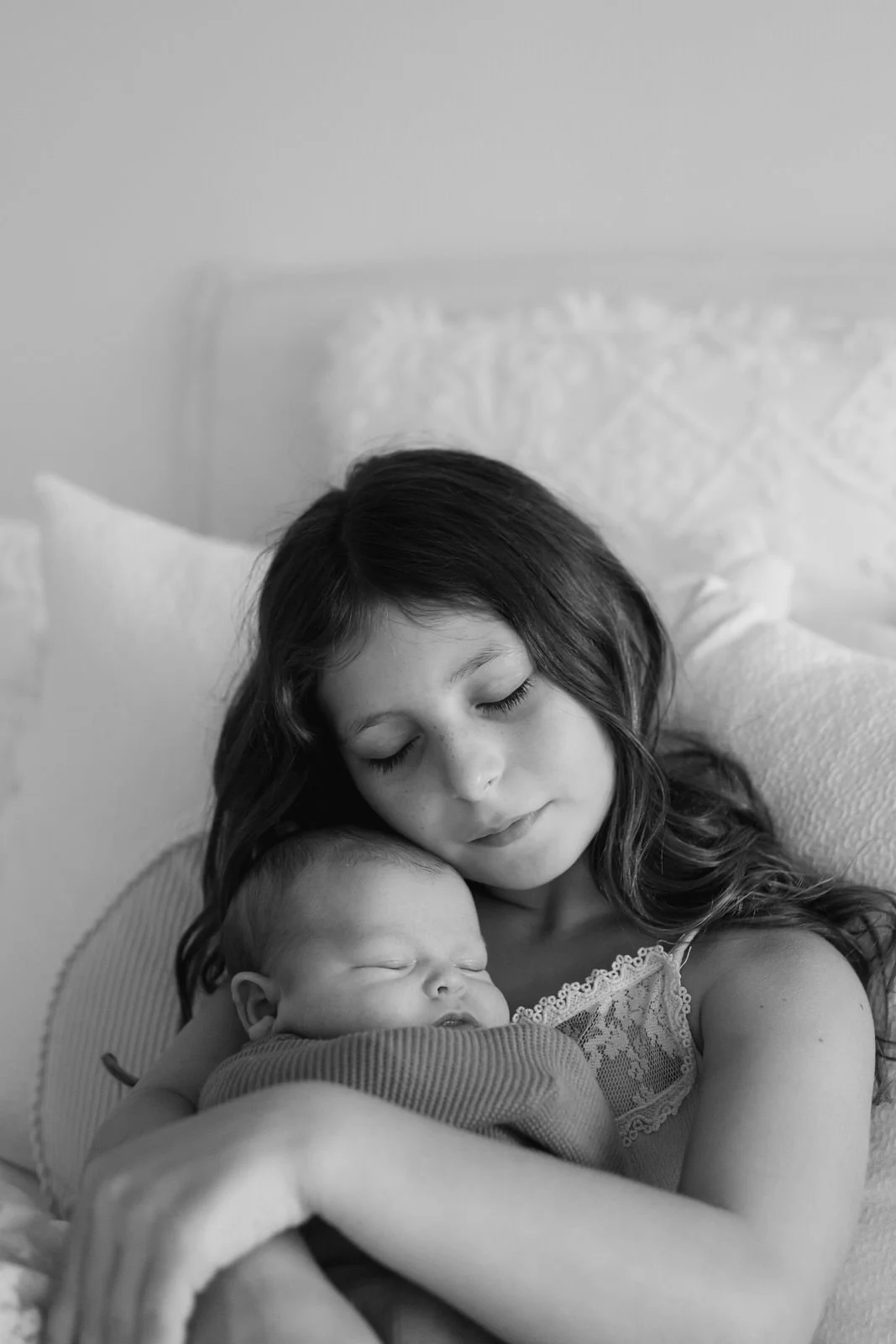 A young girl with closed eyes gently holding a sleeping infant with closed eyes, both resting on a bed with pillows, in black and white.