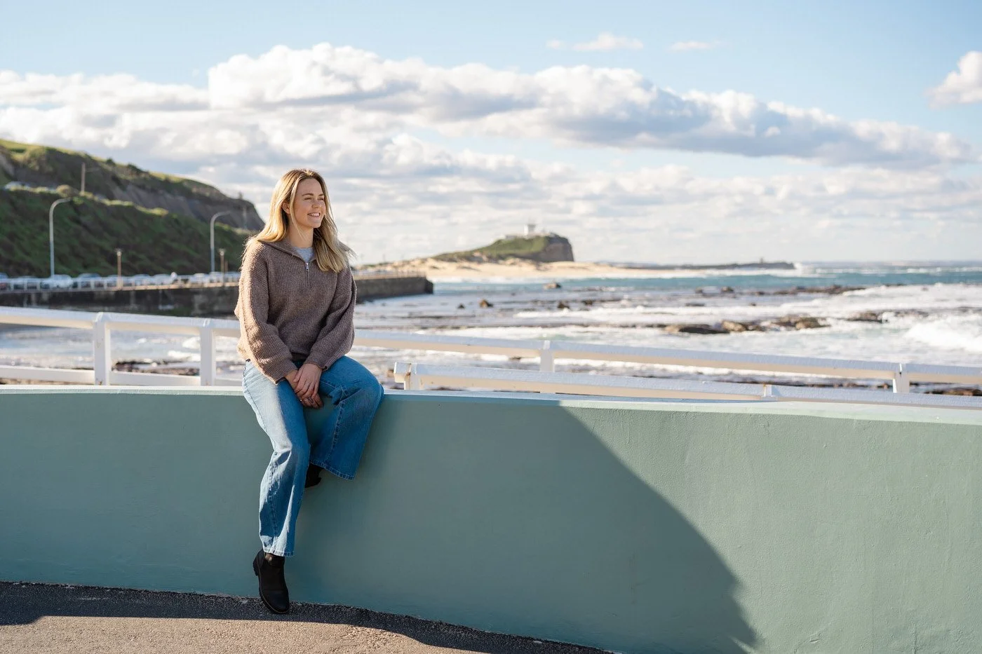 Woman sitting on a blue half wall, staring at the ocean near Nobby's beach in Newcastle NSW