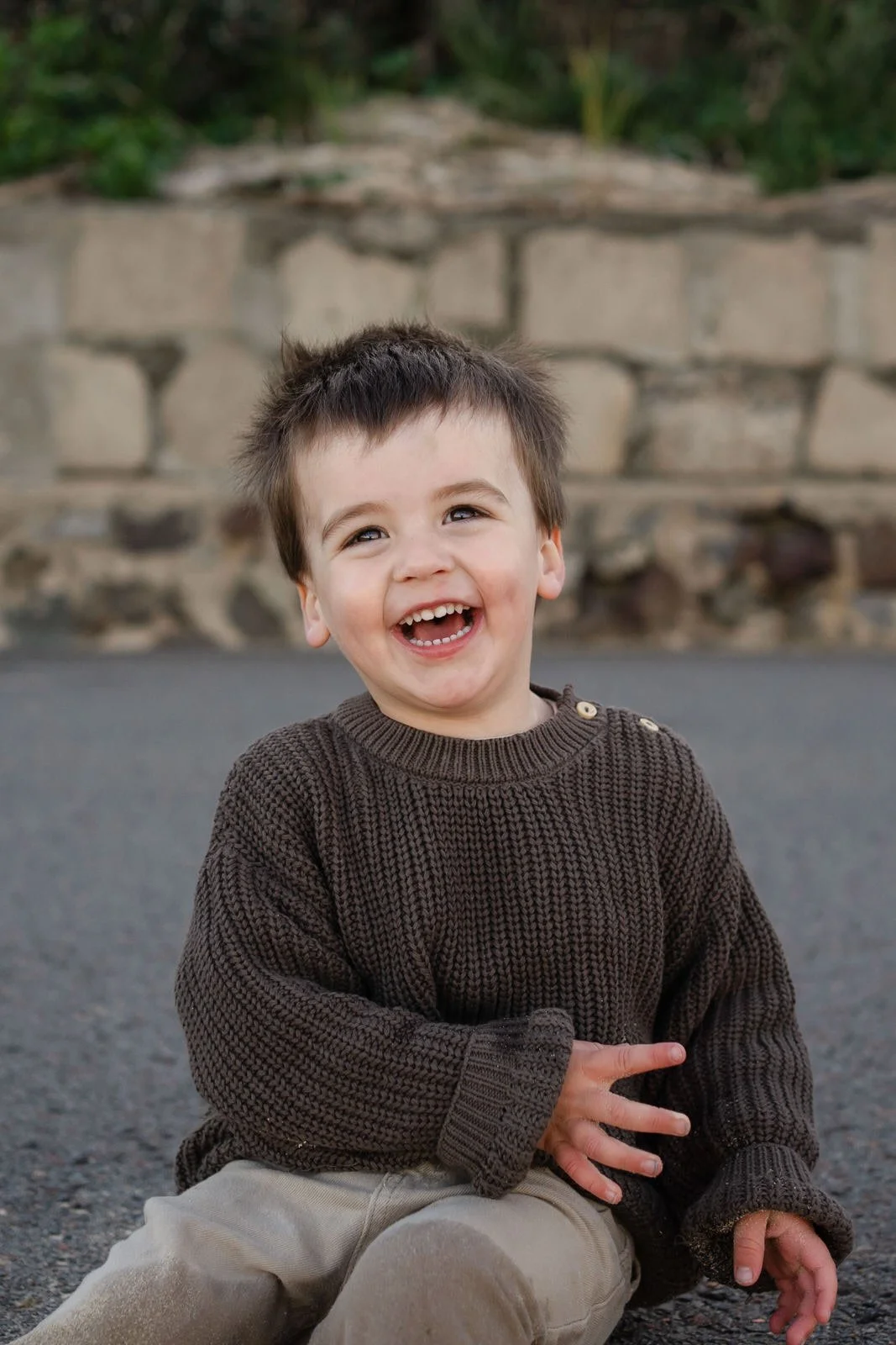 A young boy with dark hair and a big smile, sitting on the pavement outdoors, wearing a brown knit sweater and beige pants.