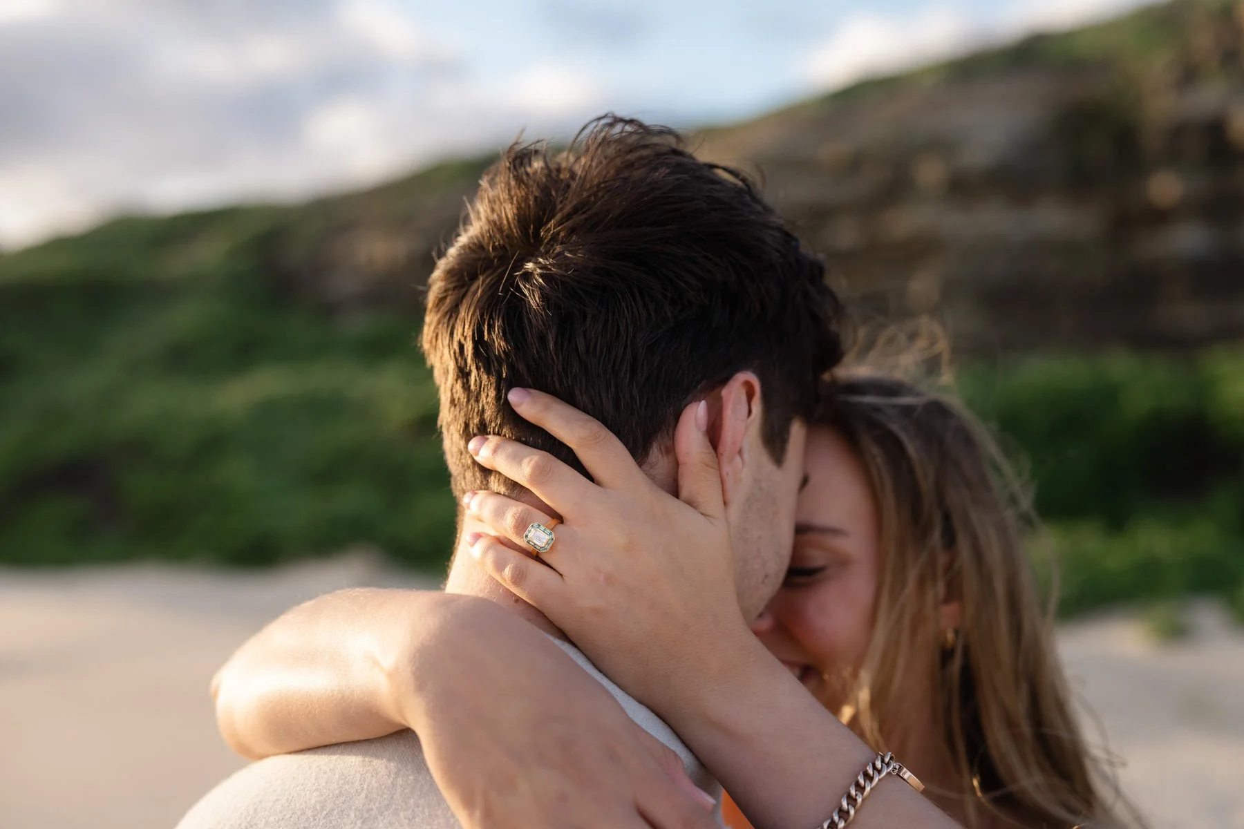 A couple embracing each other closely on a beach with green hills and cloudy sky in the background, with the woman's hand resting on the man's face, displaying a large ring.