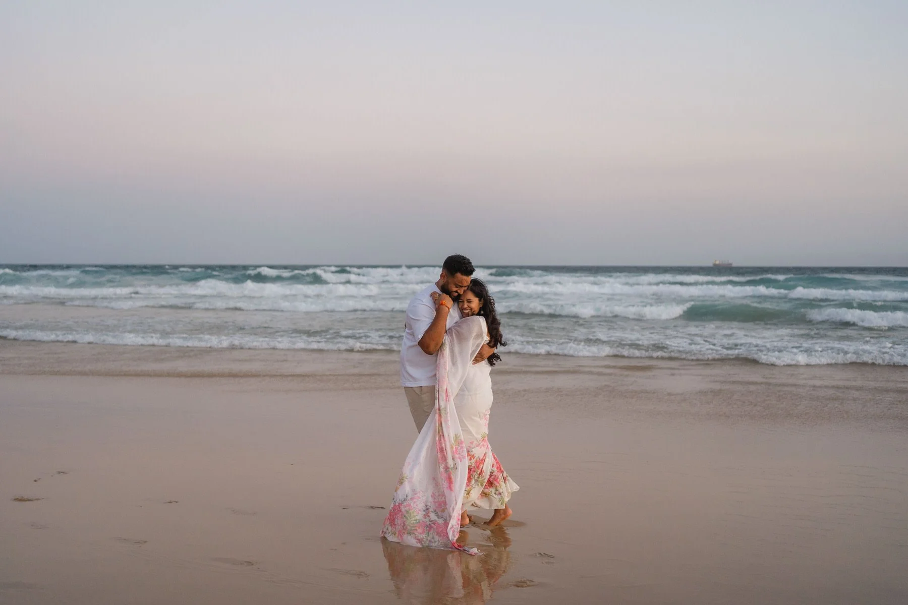 A couple hugging and smiling on the beach as the waves crash behind them during sunset or sunrise.