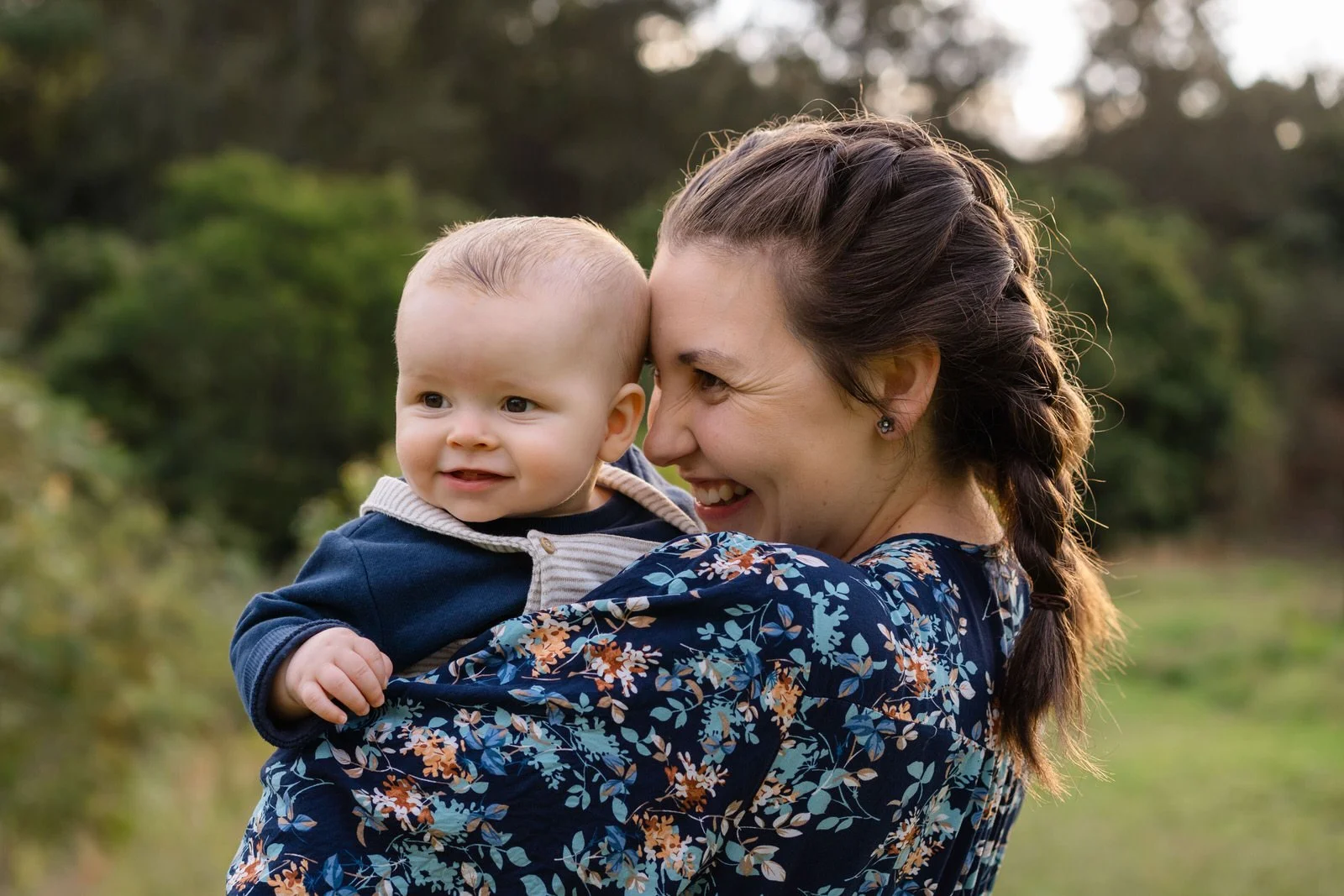 A woman with brown hair in a braid smiling while holding a baby boy outdoors with trees and greenery in the background.
