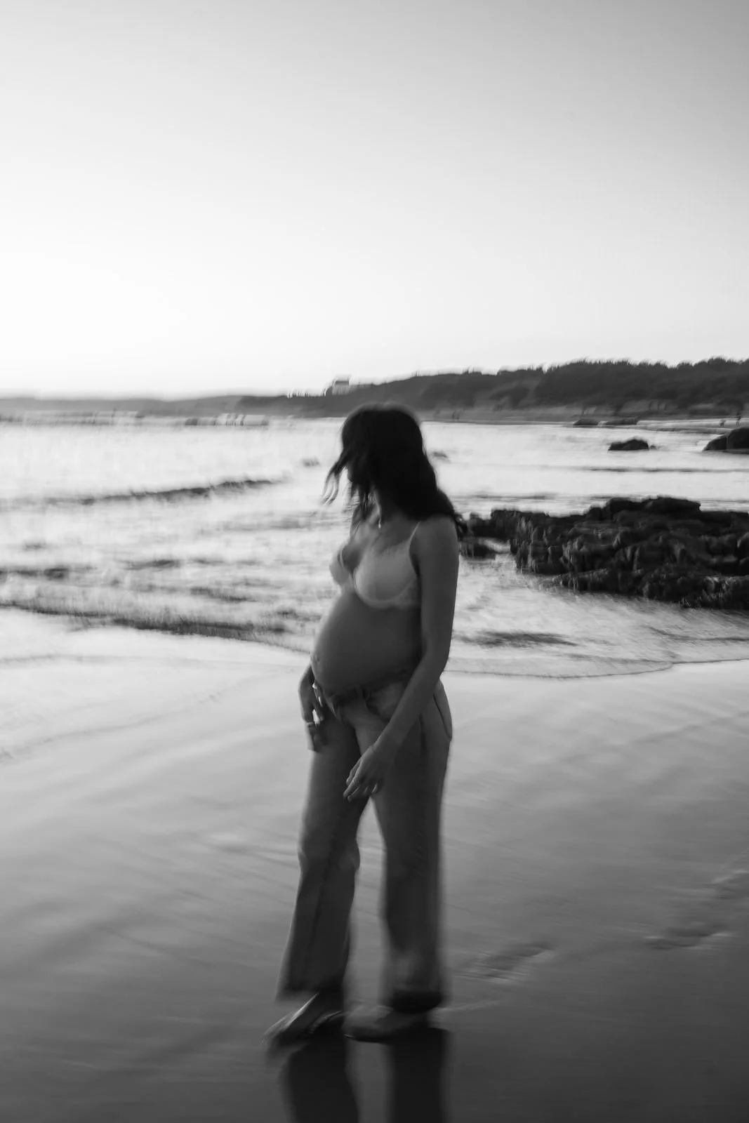 A pregnant woman in a tank top and wide-leg pants standing on a beach near the water at sunset, with rocks and distant shoreline in the background.
