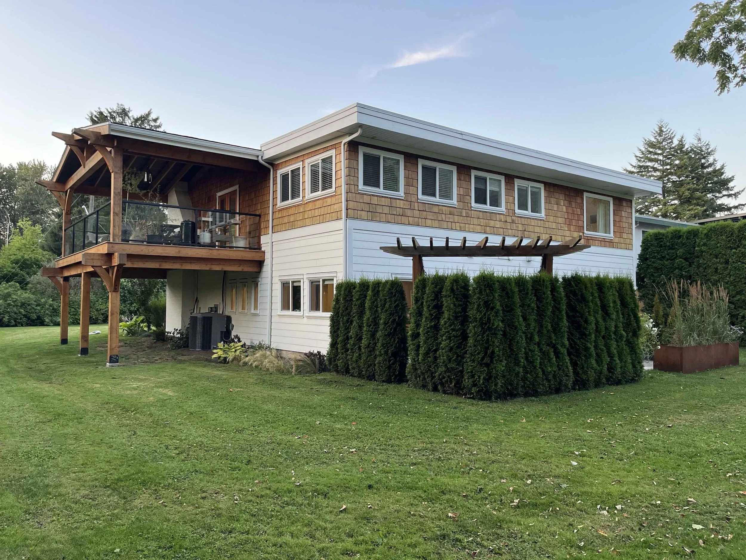 A two-story house with wooden shingles on the upper floor and white siding on the lower floor, surrounded by a well-maintained green lawn and greenery. The house features a small balcony with a black railing, and a pergola extends from the side. Tall trees are visible in the background under a partly cloudy sky.