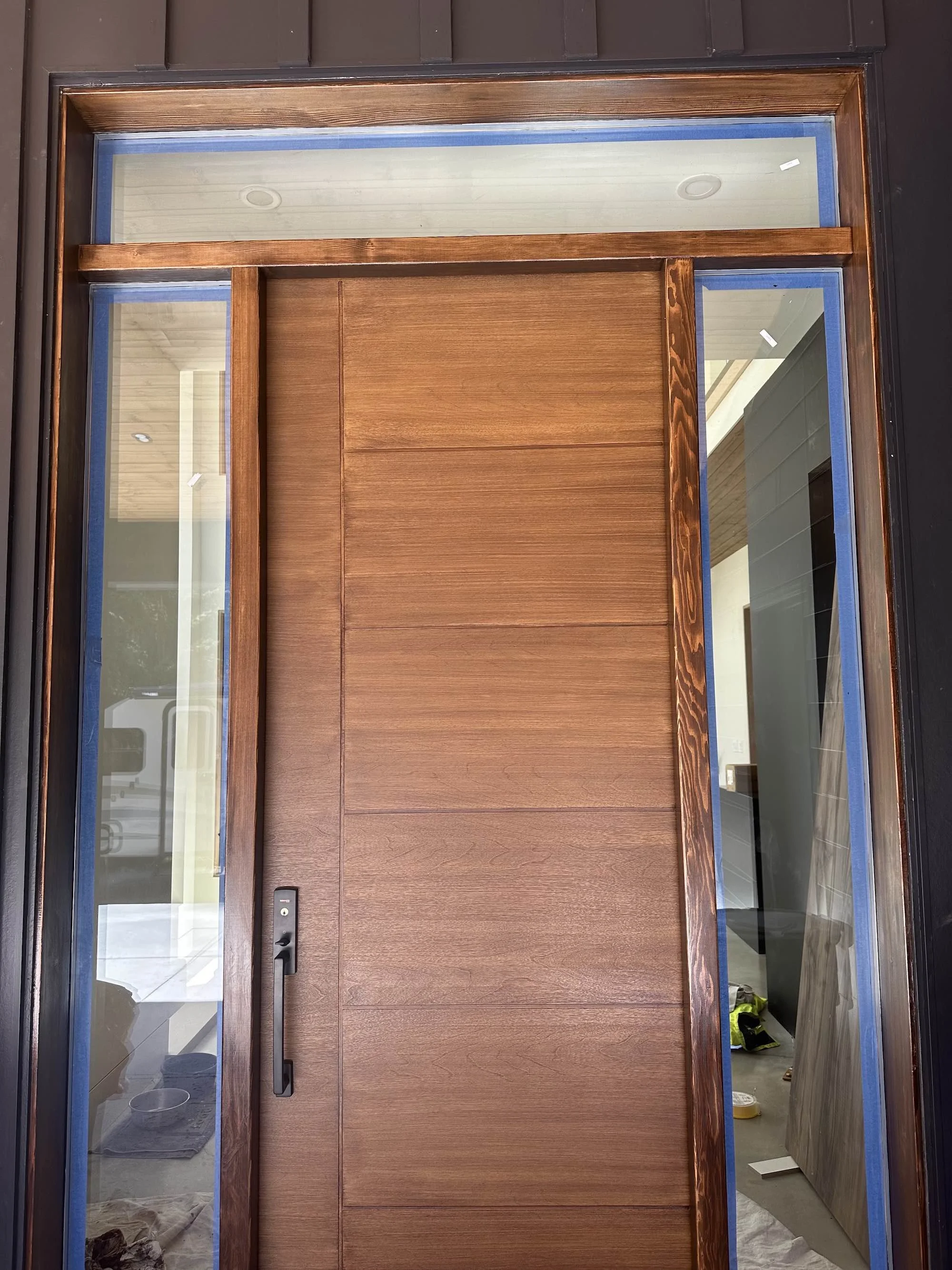Wooden front door with glass panels on each side and above, black exterior frame, door handle, visible interior room with construction supplies and tools.