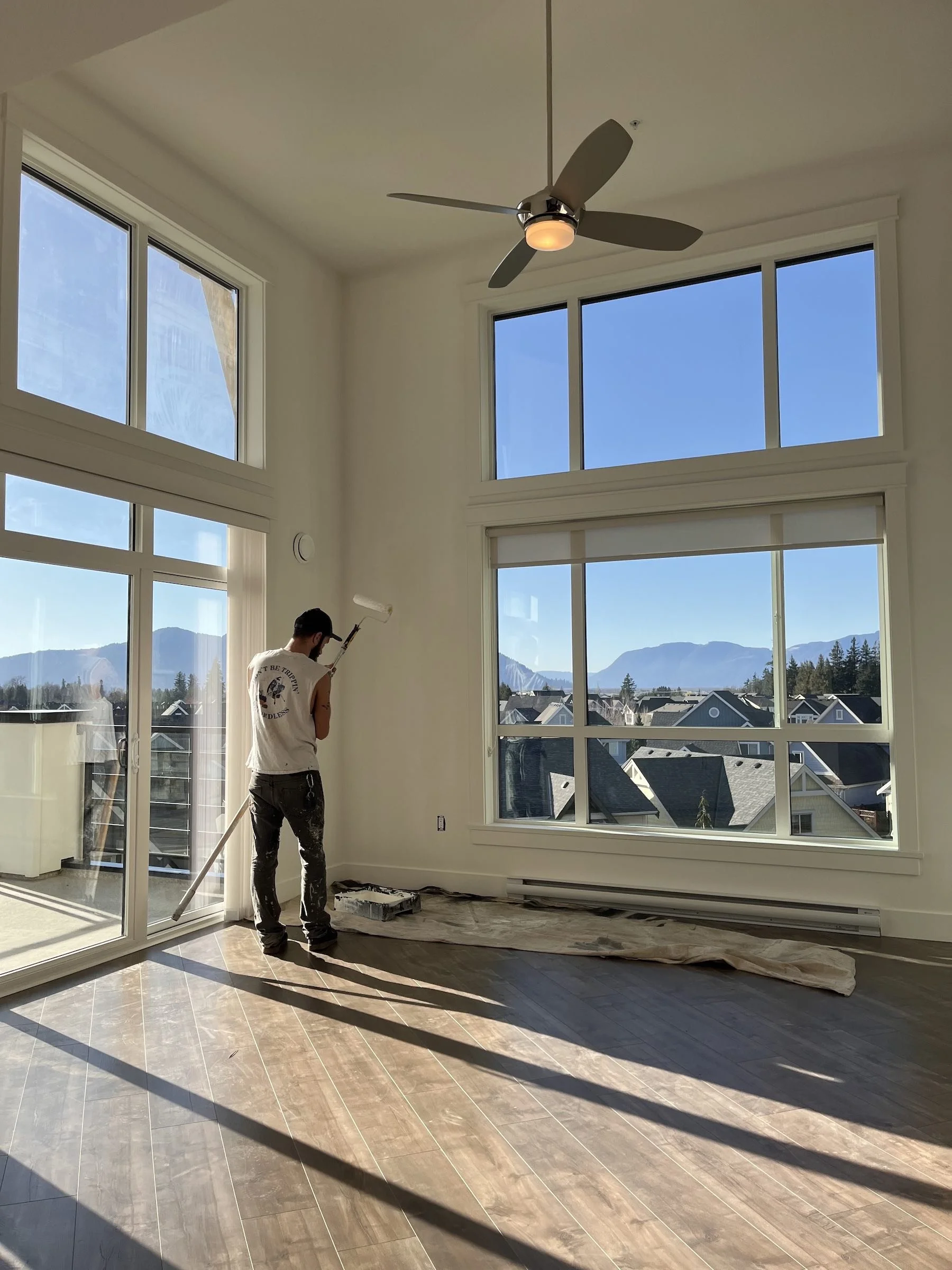 Person painting walls inside a room with large windows, view of mountains and houses outside, sunlight streaming in.