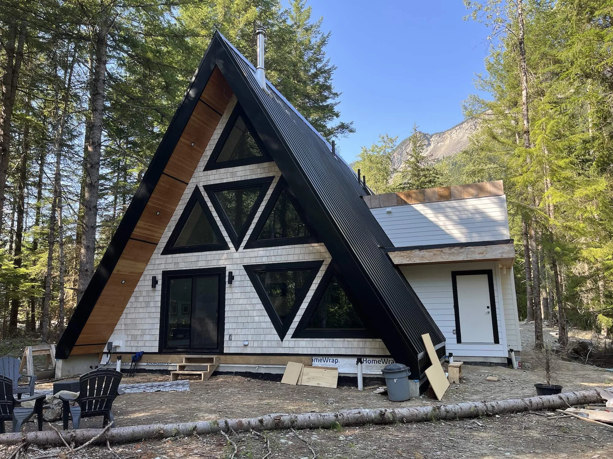 A modern A-frame house under construction in a wooded area with tall trees and mountains in the background. The house has black triangular windows, a mix of white and wood exterior siding, and construction materials and debris are visible around the site.