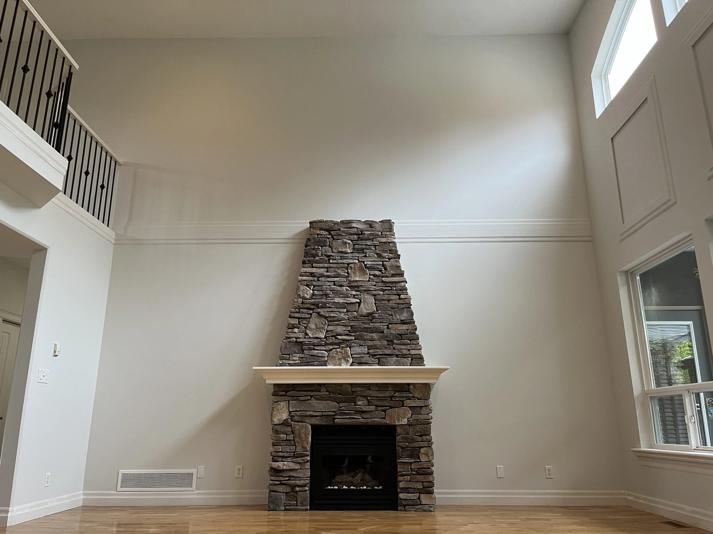 Living room with a rustic stone fireplace, high ceiling, large windows, white walls, hardwood floor, and a side railing on the upper level.