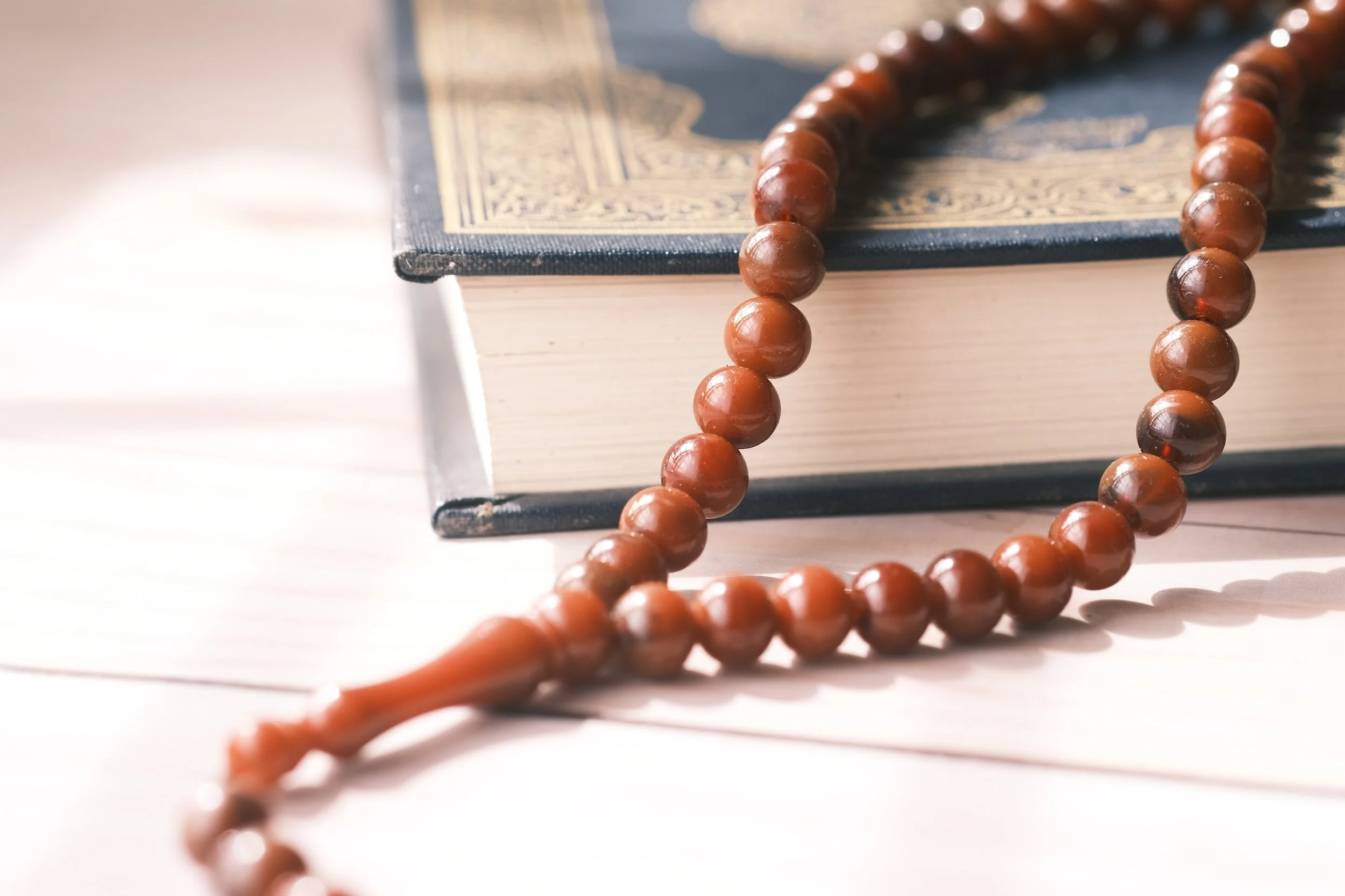 A string of wooden prayer beads resting on a white wooden surface, with a closed book with dark cover and ornate gold details in the background.