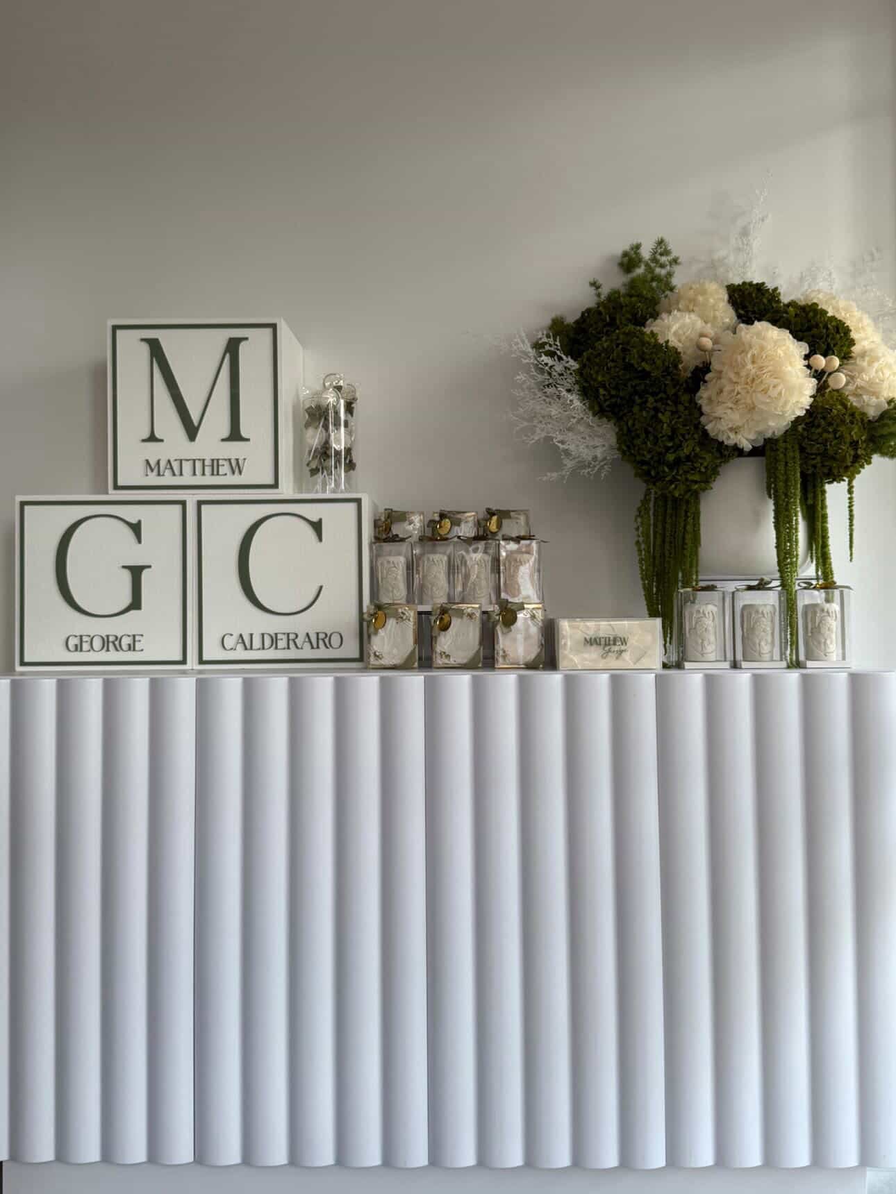 Decorative display with white boxes featuring initials and names, surrounded by silver gifts, a floral arrangement of white and green flowers, and a table covered with white textured fabric.