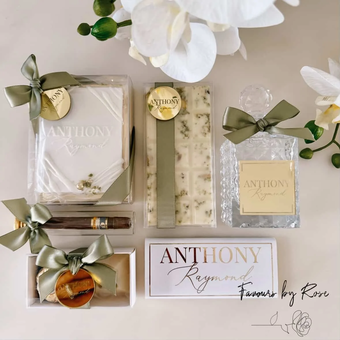 Gift set with wrapped soaps, a candle, and a perfume bottle, all labeled 'Anthony Raymond,' decorated with green ribbons, arranged on a table with white orchids.