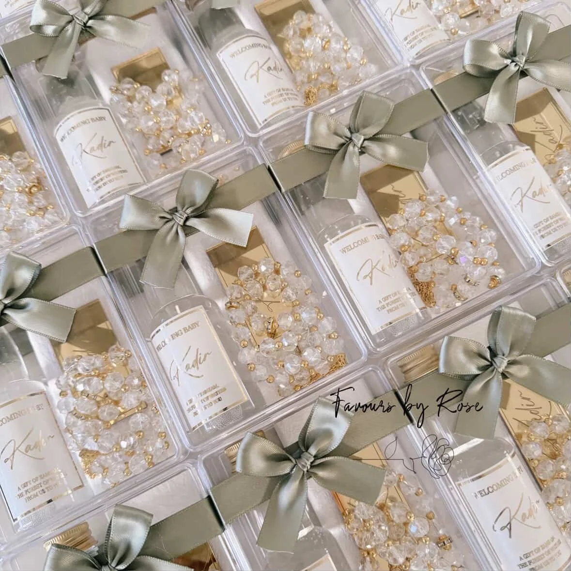 Clear plastic boxes tied with olive green ribbons, containing pearl and gold bead jewelry, labeled "Favors by Rose" for a baby shower.