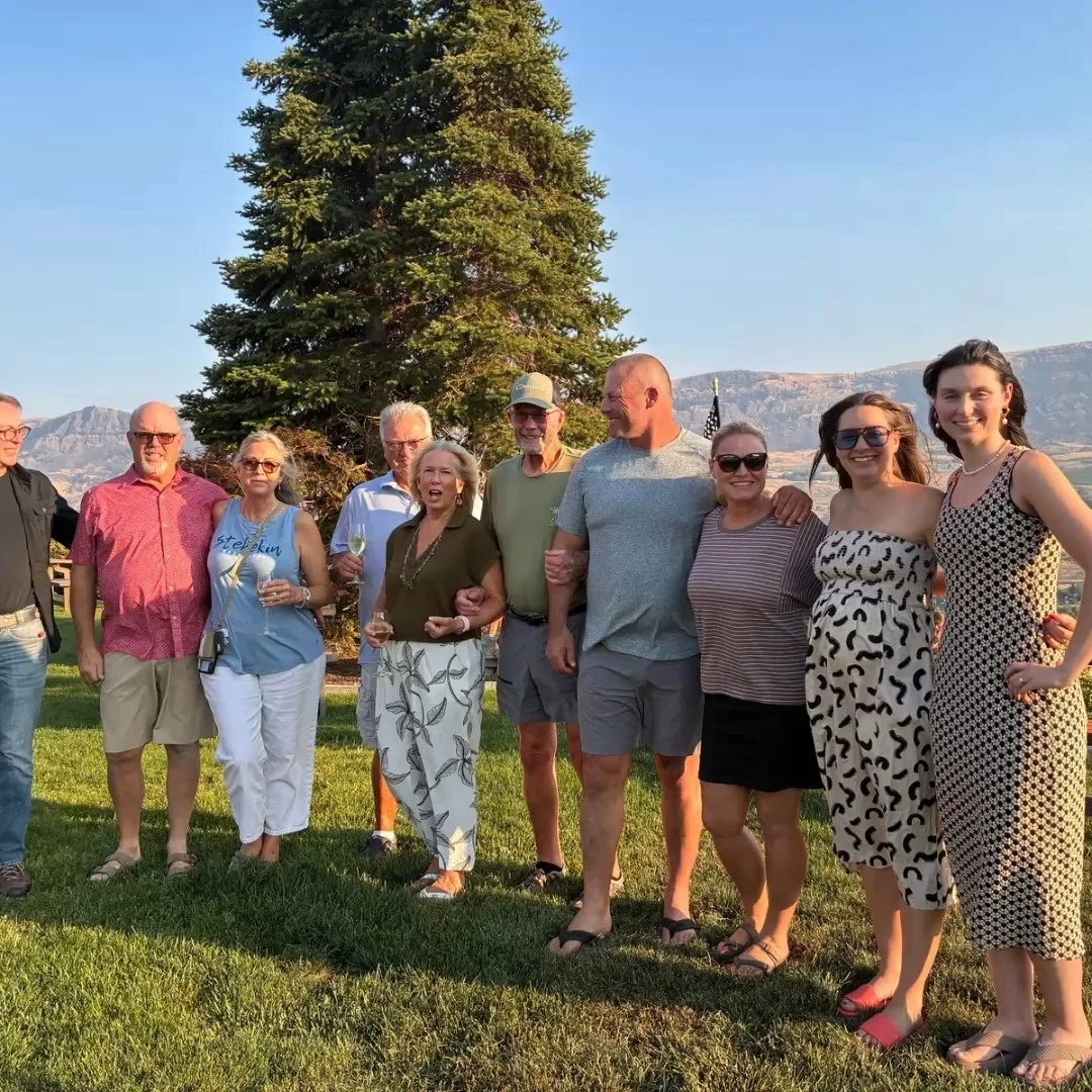 A group of people standing outdoors on a grassy lawn, with a large tree and mountains in the background, enjoying a sunny day.