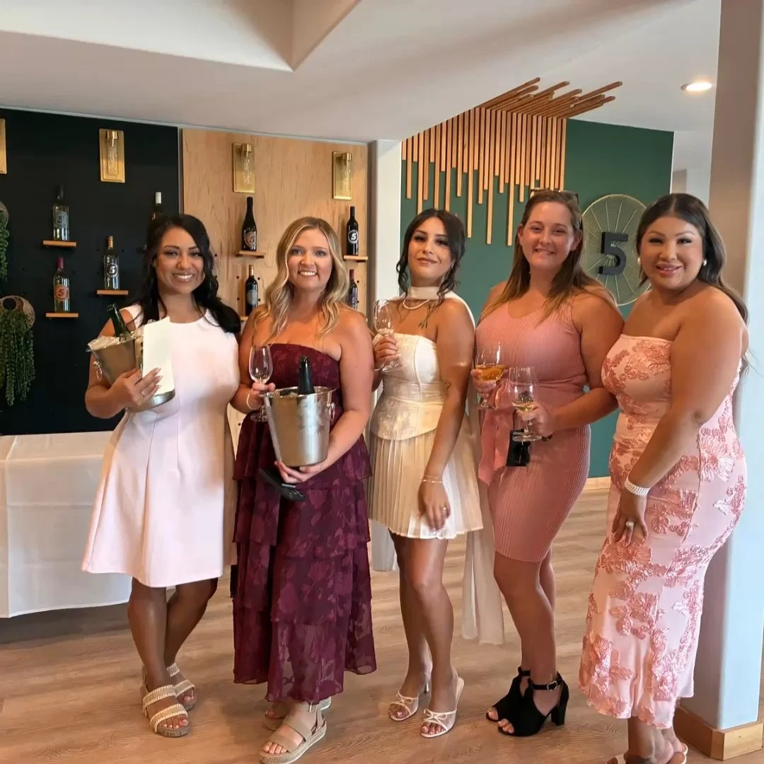 Six women in elegant dresses holding wine glasses posing for a photo at a party or celebration in a modern indoor setting.