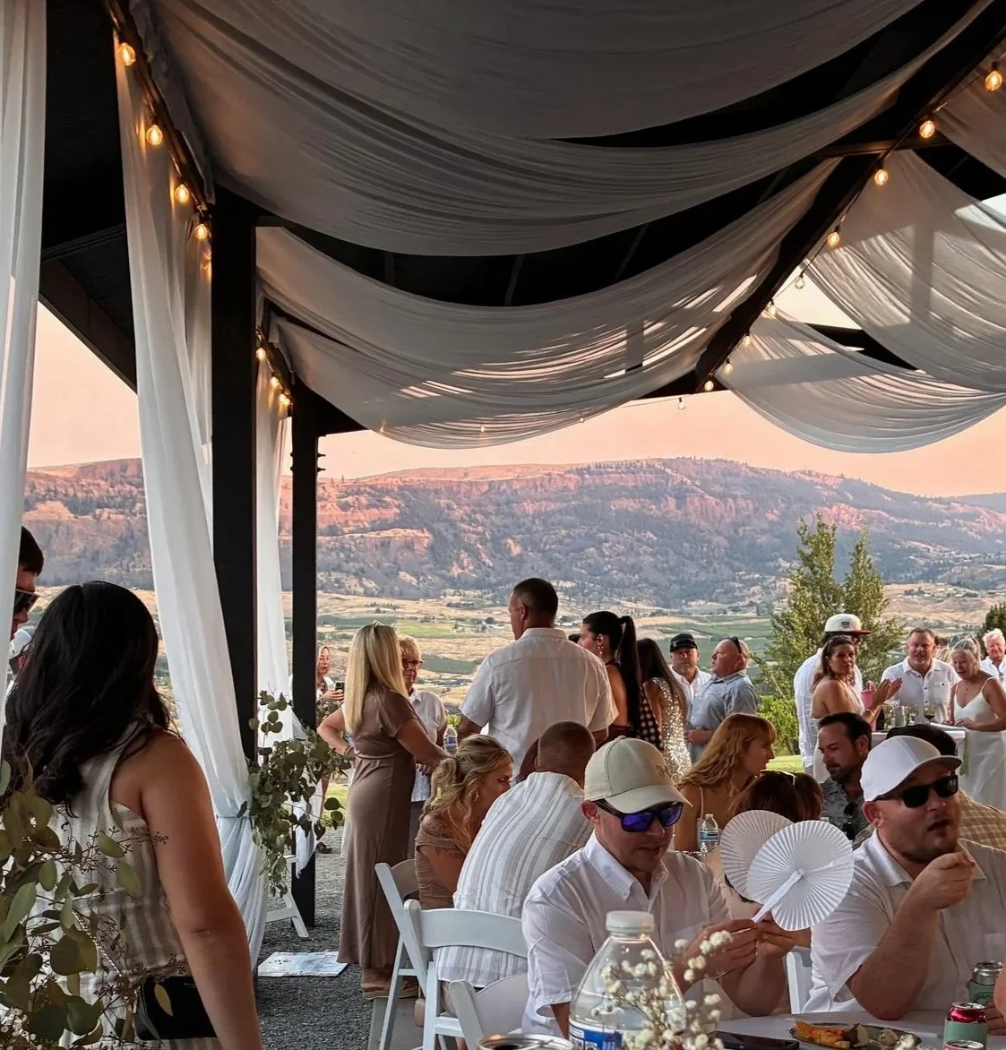 People gathered at an outdoor event with mountains in the background, under a decorated tent with draped fabric and string lights.