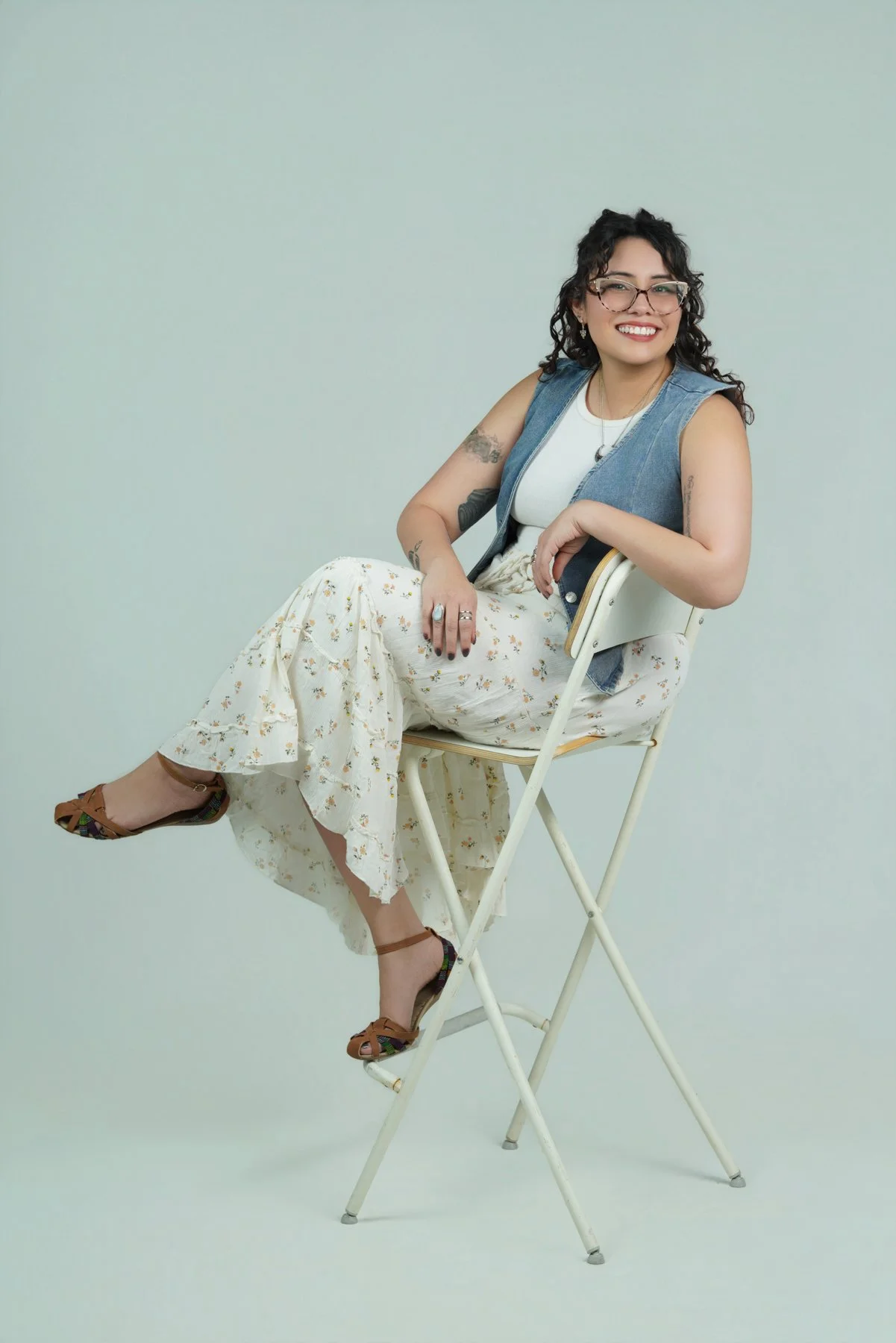 image of tattooed therapist in los angeles with curly hair wearing glasses, floral skirt, denim vest sitting on chair