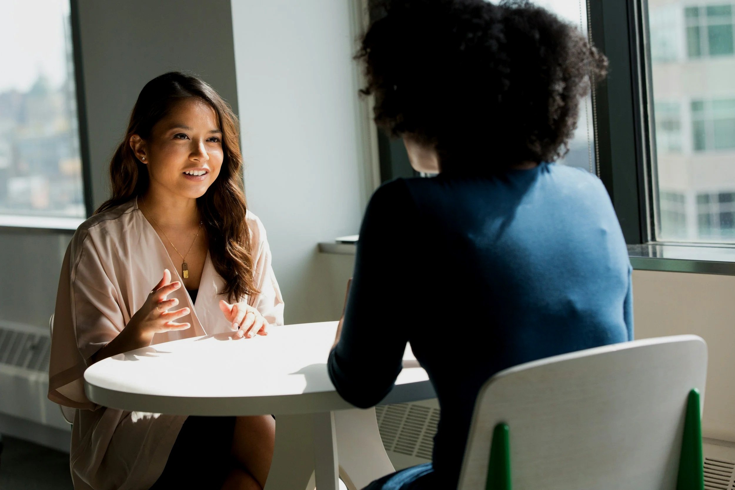 two diverse women sitting across from each by a window facing each other talking