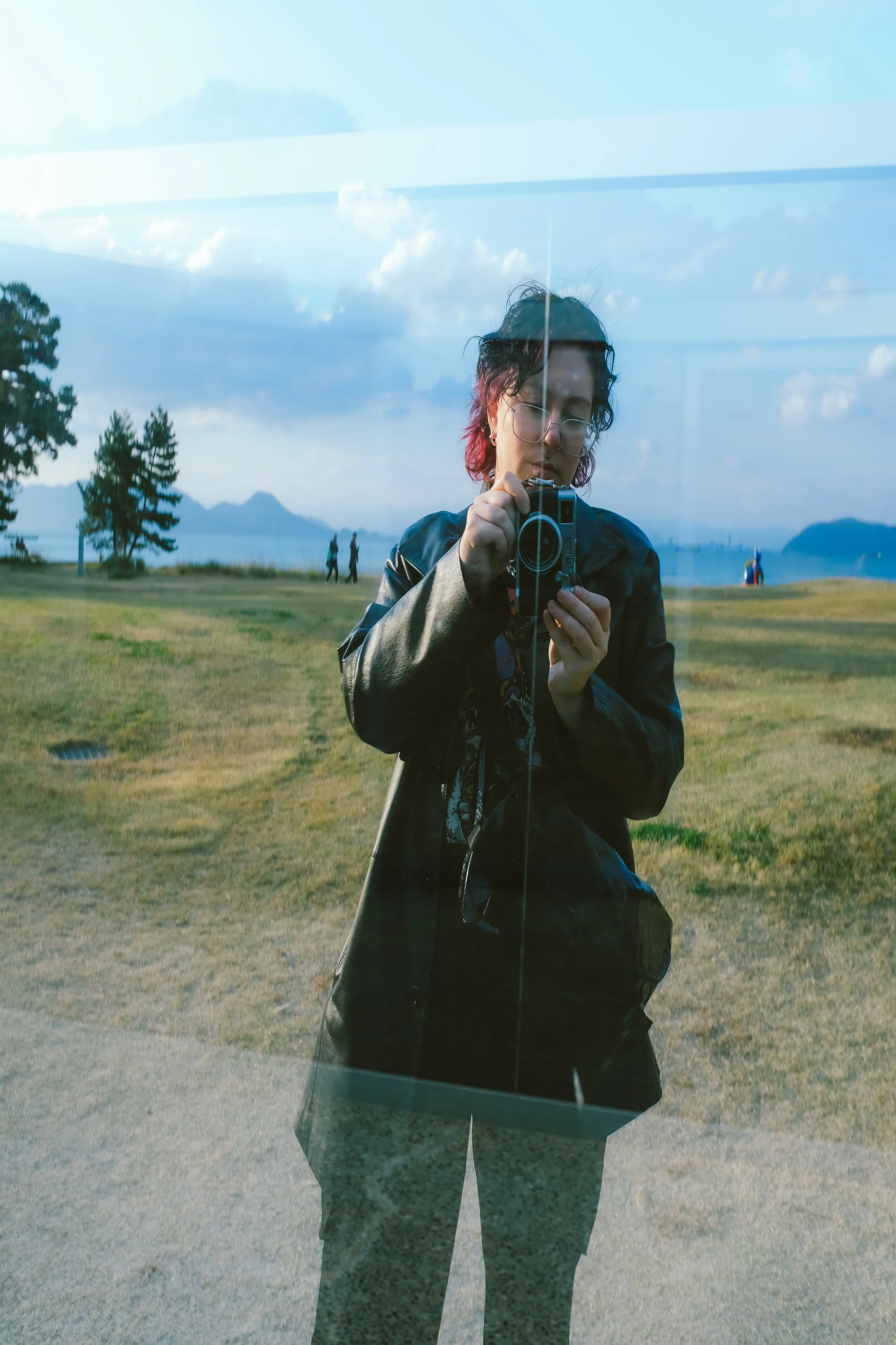 A person taking a photo of their reflection on a glass window, with a landscape of grassy field, trees, and mountains in the background.