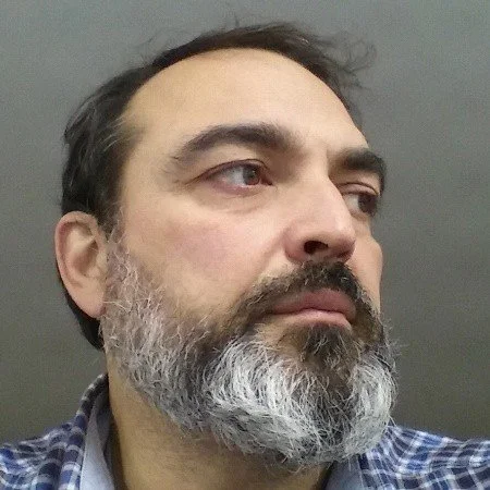 Close-up of a man with a beard and mustache, wearing a plaid shirt, looking to the right against a gray background.