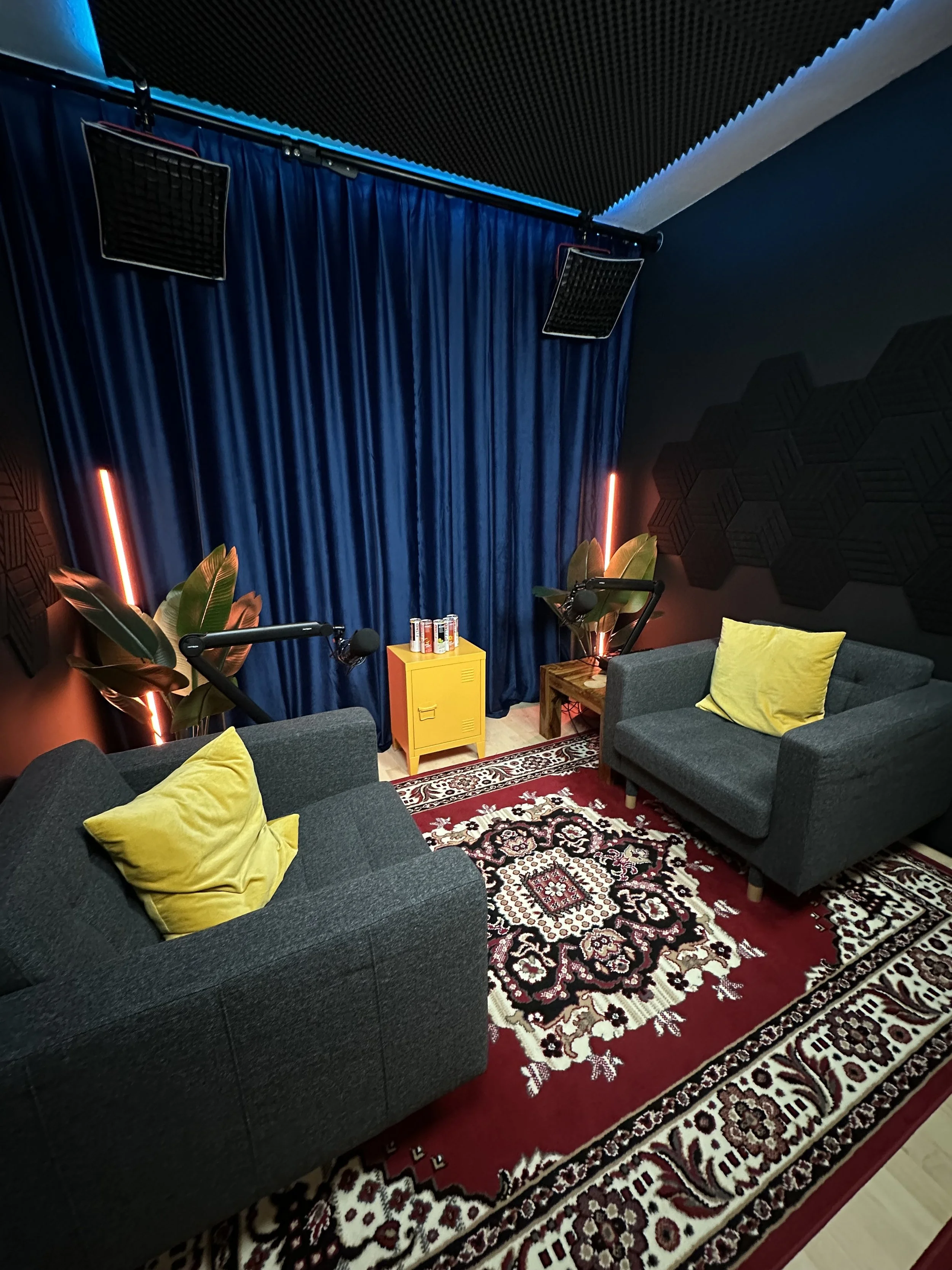 A cozy soundproof recording or podcasting studio with two dark gray couches with yellow pillows, a patterned area rug, a small yellow cabinet with drinks, two large plants, blue curtains, black acoustic foam panels, and two mounted lights.
