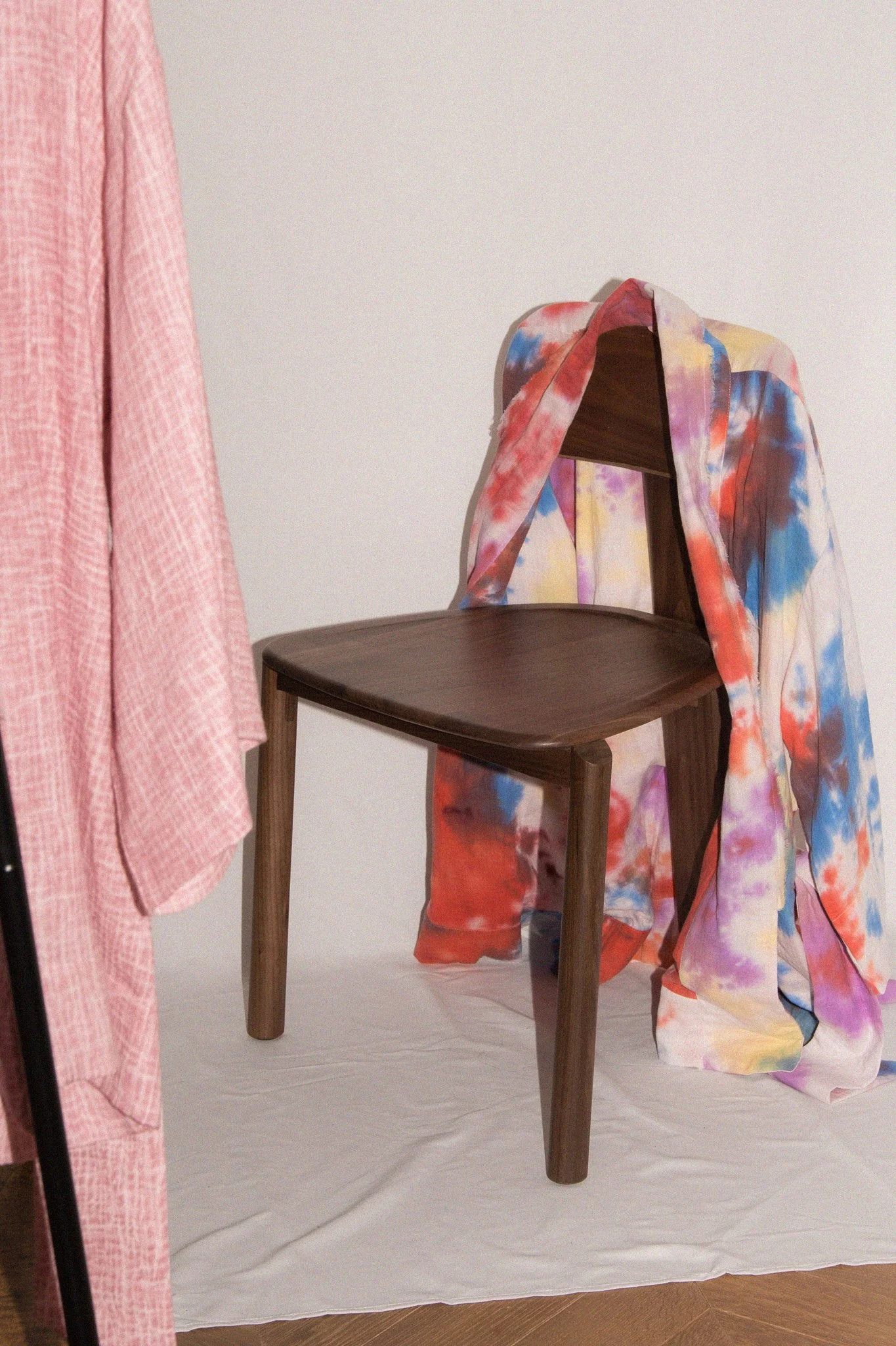 Une chaise en bois foncé avec un tissu coloré en arrière-plan et un vêtement rose suspendu à gauche.