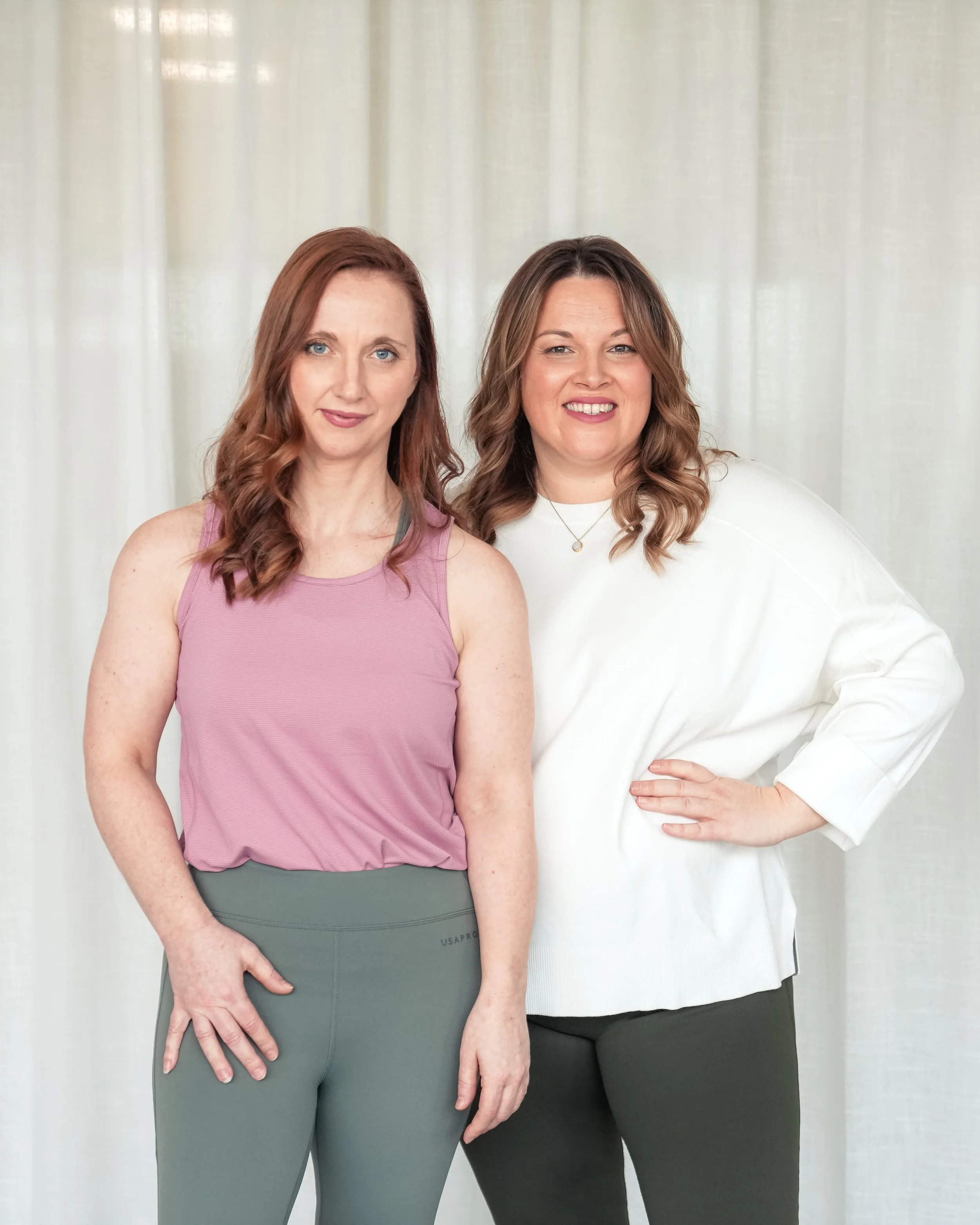 The two female Thrive Studio owners standing in front of a light-colored curtain, smiling at the camera.