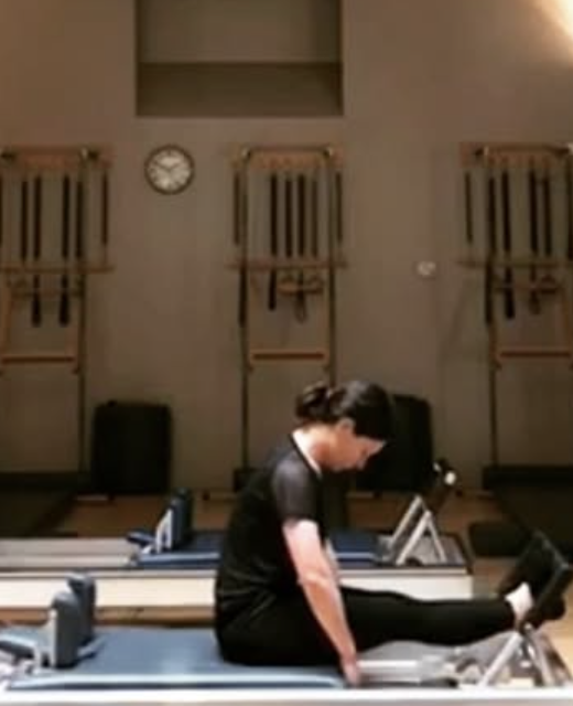 A woman practicing yoga, balancing on a yoga block with her body turned sideways, her arms extended overhead in a curved position, with her eyes closed.