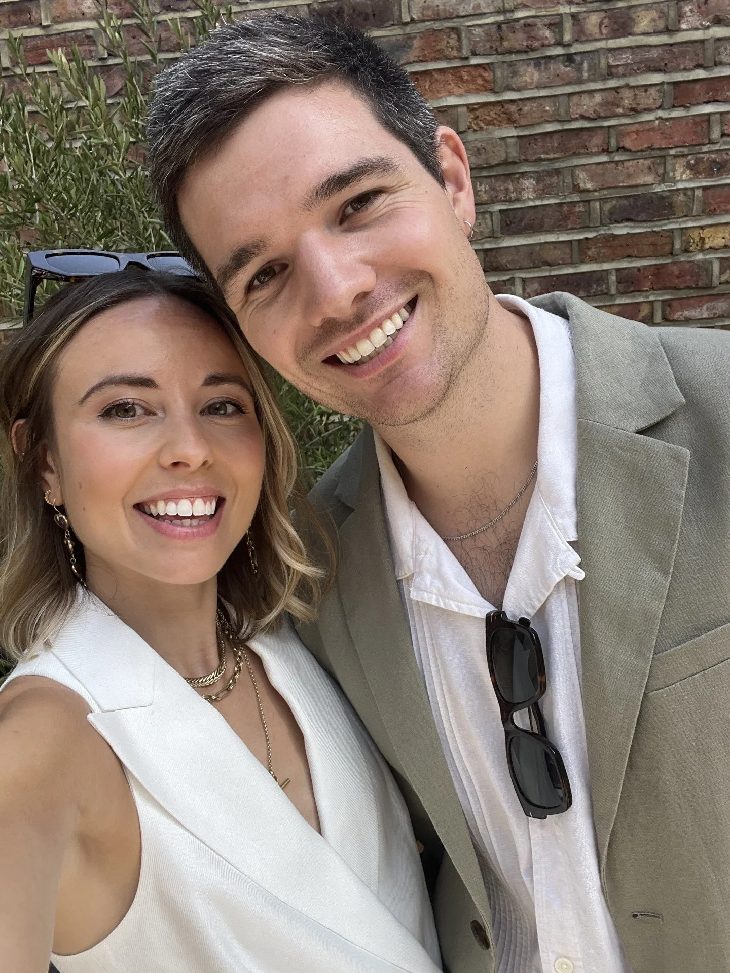 Smiling person wearing a white 'The Good Life' T-shirt with a tree and sun design, standing outdoors in front of iron fence and trees.