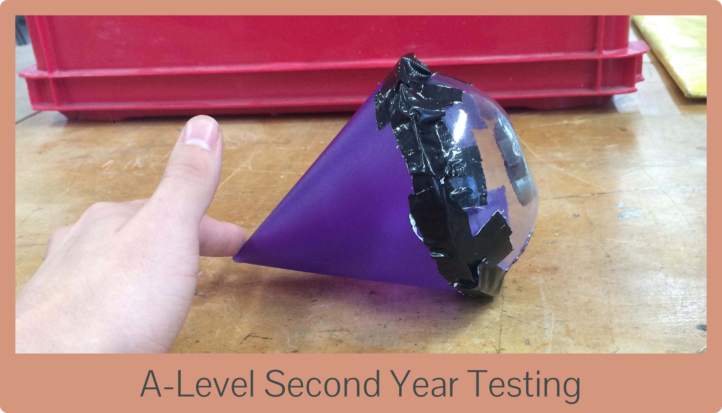 A hand holding a purple cone-shaped object with black tape on one end, placed on a wooden table with a red plastic container in the background.