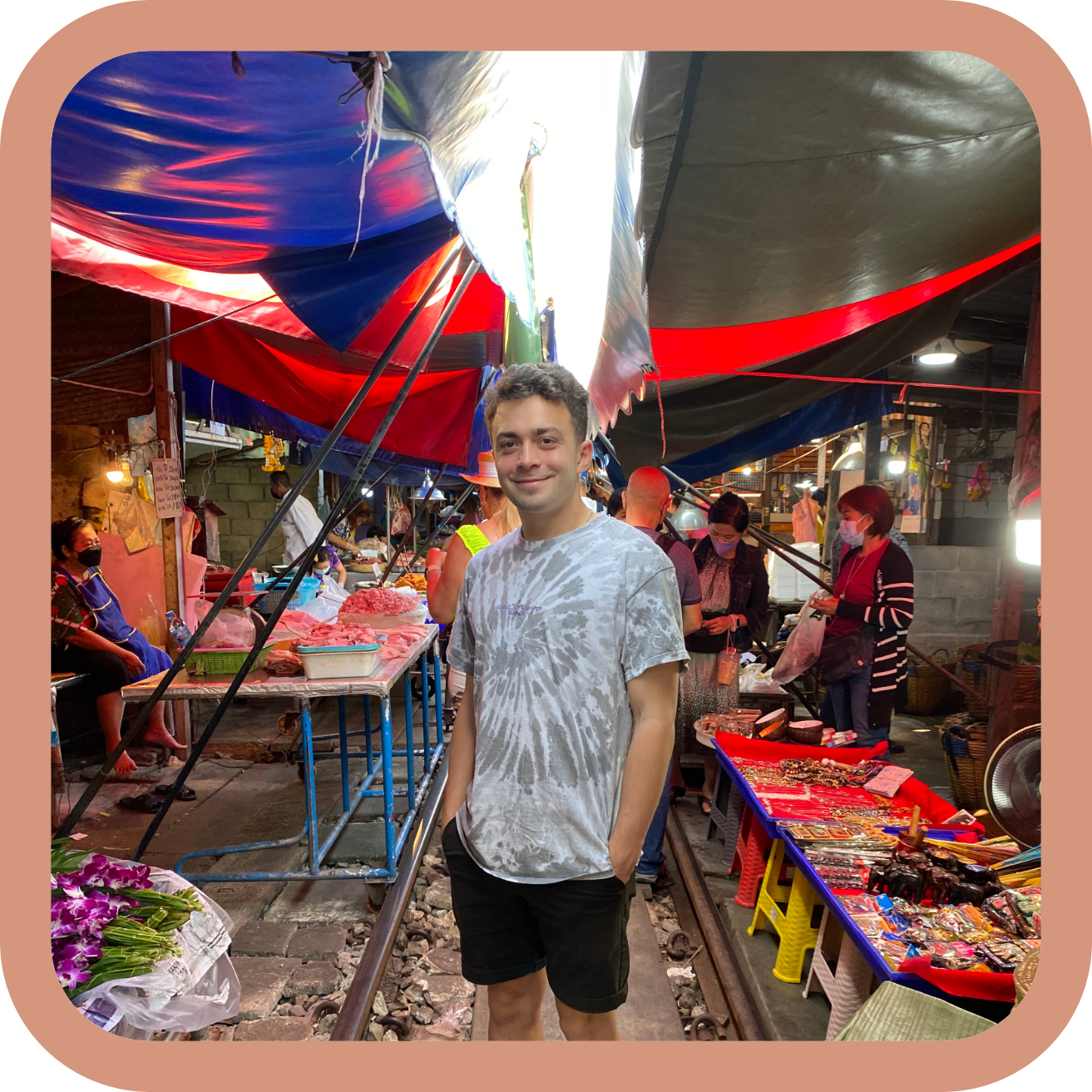 A young man in a tie-dye t-shirt and black shorts standing at a bustling outdoor market with colorful umbrellas, vendors, and shoppers in the background.