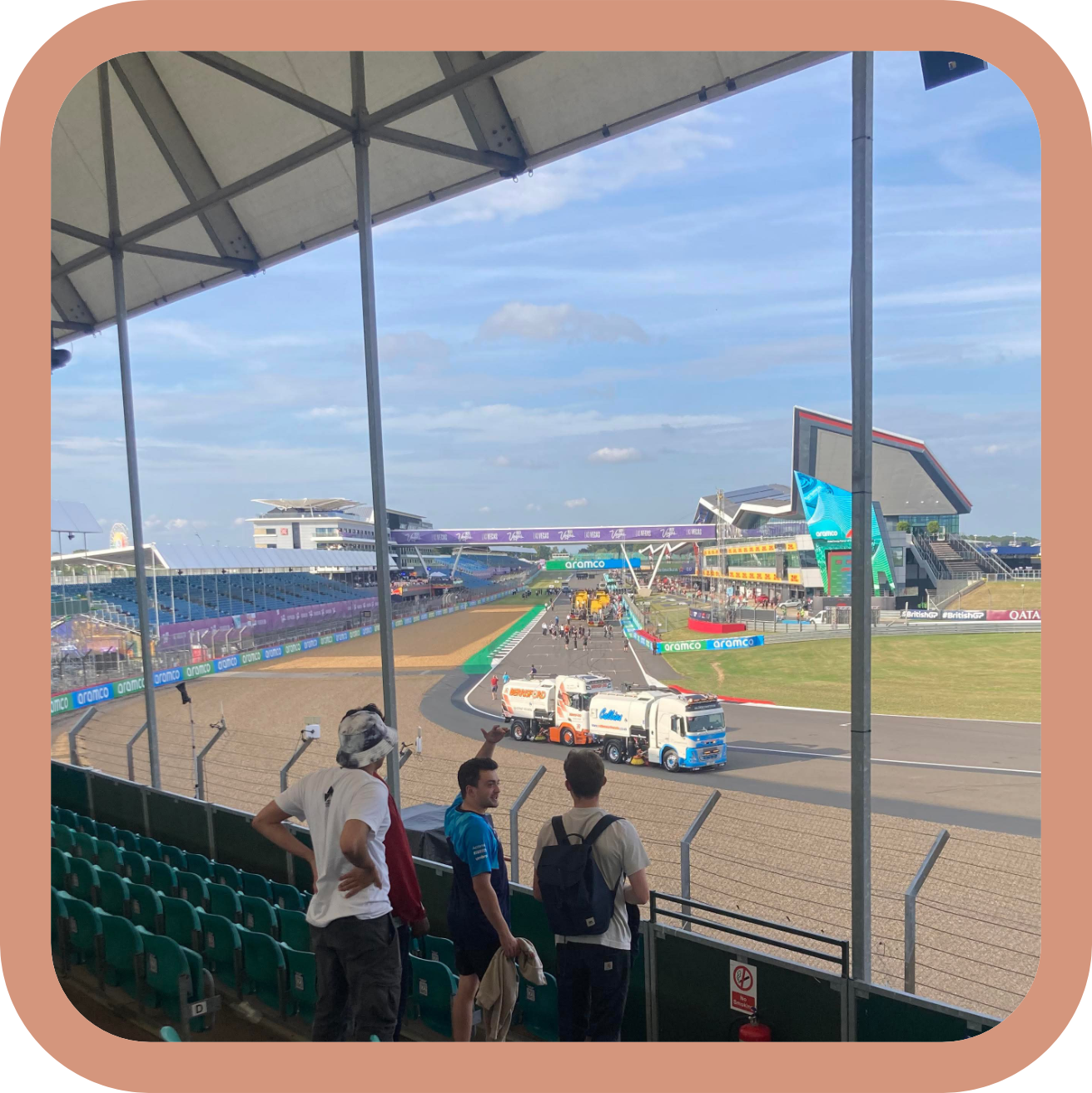 View of a race track with trucks and spectators, under a partly cloudy sky, with grandstands and modern buildings in the background, as seen from a covered seating area.