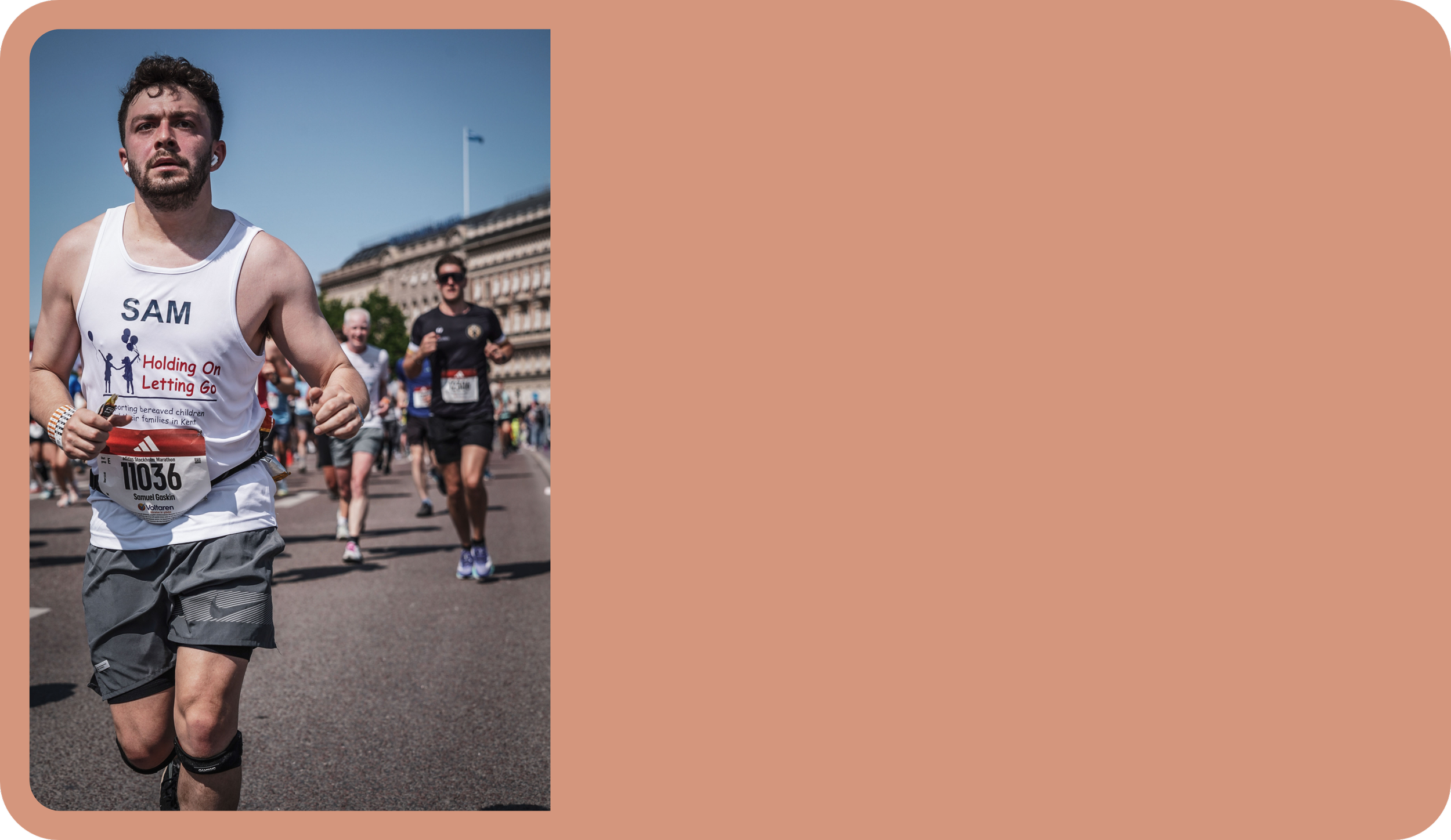 A man wearing a white tank top with "SAM" on it and gray shorts runs in a marathon on a city street with other runners behind him, along with spectators and historic buildings.