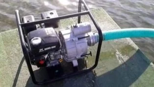 A small water pump mounted on a metal frame, connected to a flexible hose, placed on a floating surface on water.