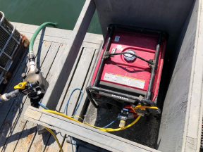 Photo of a red marine battery inside a black waterproof container on a boat deck, connected to wires and a microphone.