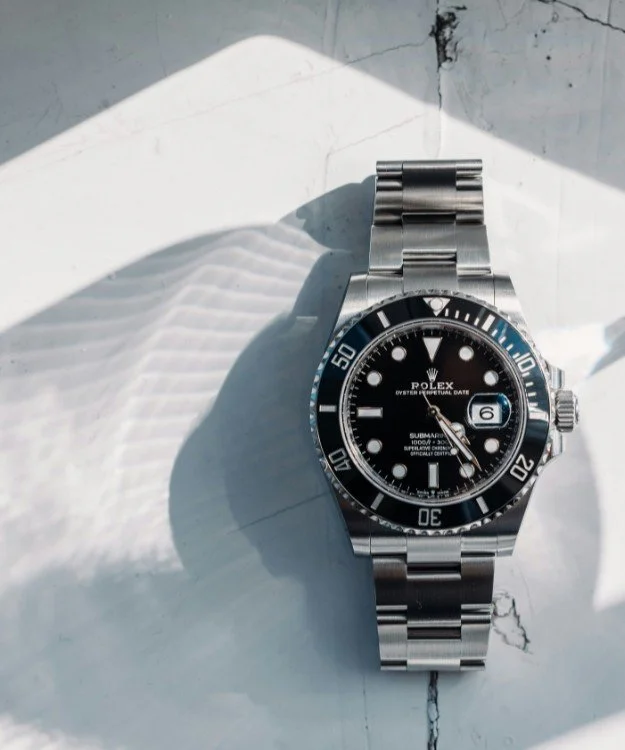 A silver Rolex Submariner watch with a black dial and bezel, positioned on a white marble surface with shadows and light reflections.