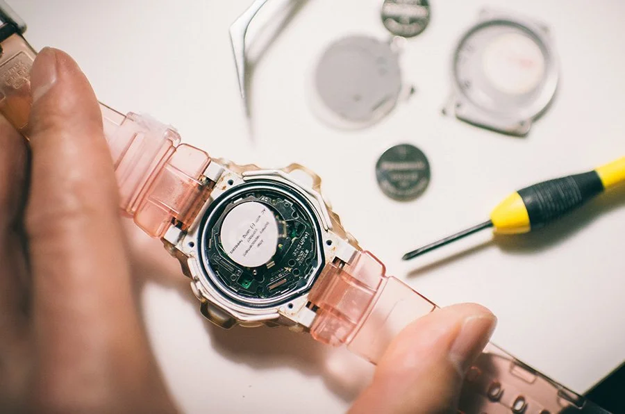 A person repairing a digital wristwatch with its back removed, surrounded by small tools and watch batteries on a white surface.