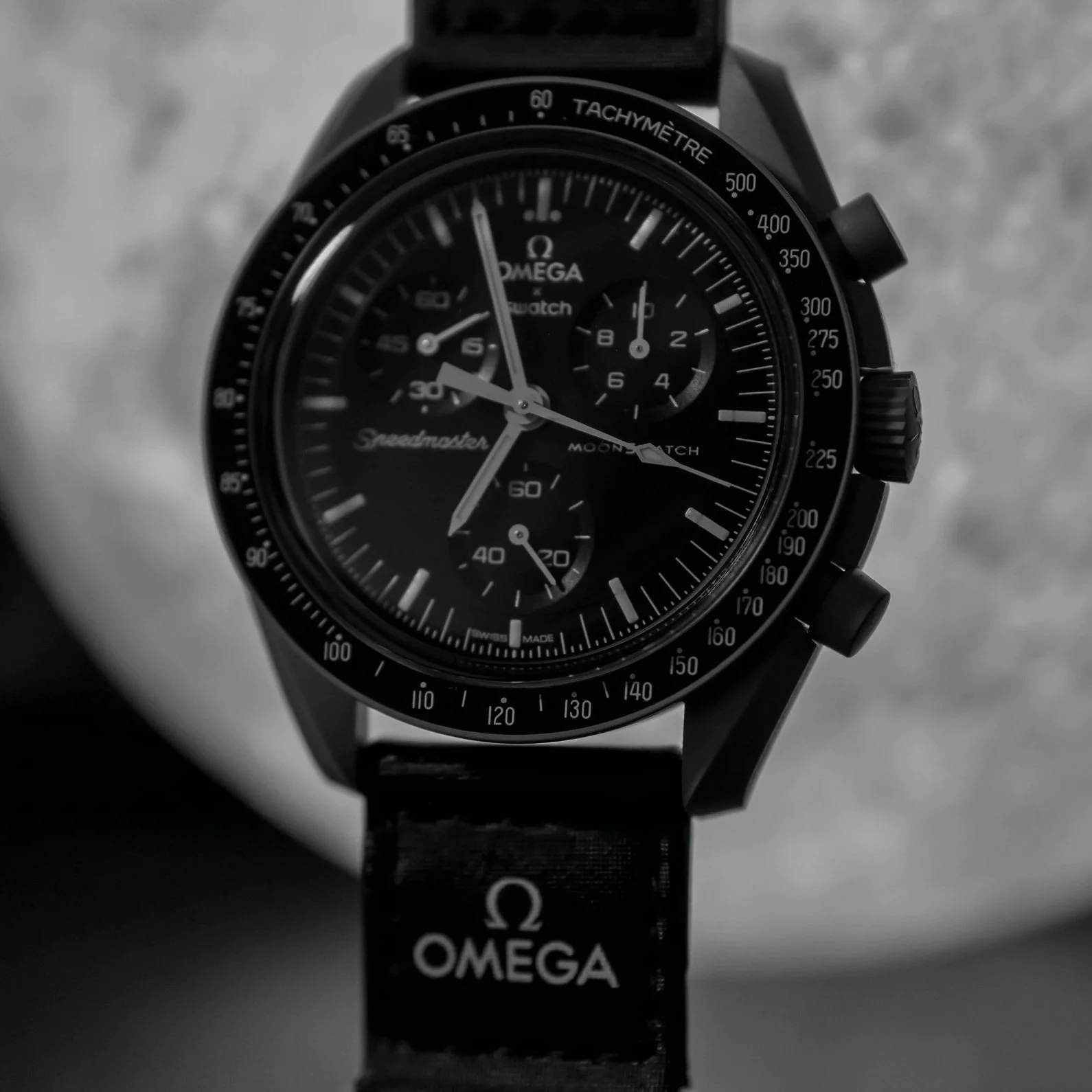 A black omega Speedmaster wristwatch with a chronograph, tachymeter bezel, and multiple small dials on a black strap, against a blurred background.