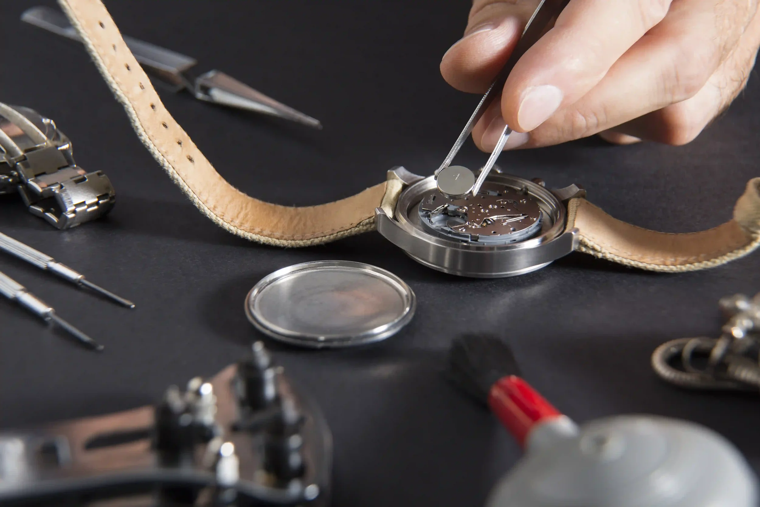 A person repairing a digital wristwatch with its back removed, surrounded by small tools and watch batteries on a white surface.