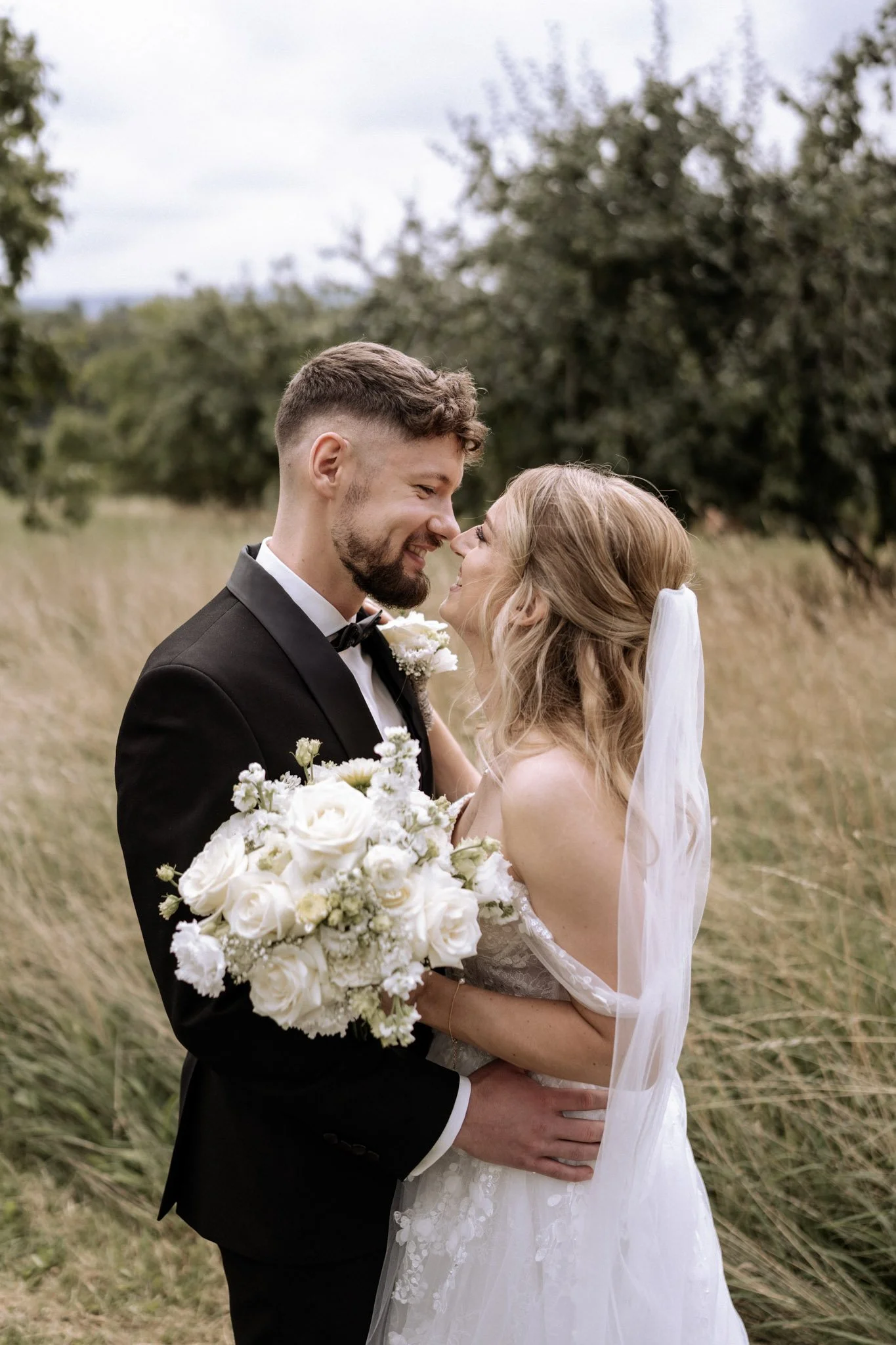 Ein Brautpaar bei einer Hochzeit im Freien, das sich liebevoll berührt und aufeinander zuneigt, die Braut hält einen großen Bouquet aus weißen Rosen, während der Bräutigam ein schwarzes Anzug trägt.