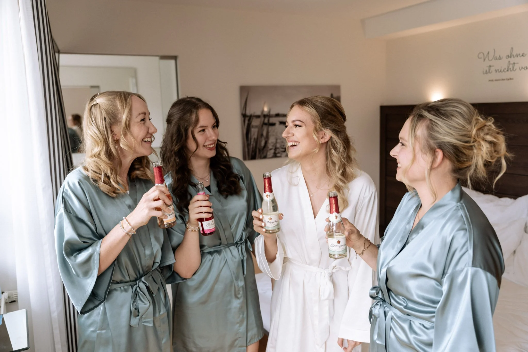 Vier Frauen in Bademänteln, lachen und trinken Bier in einem Hotelzimmer.