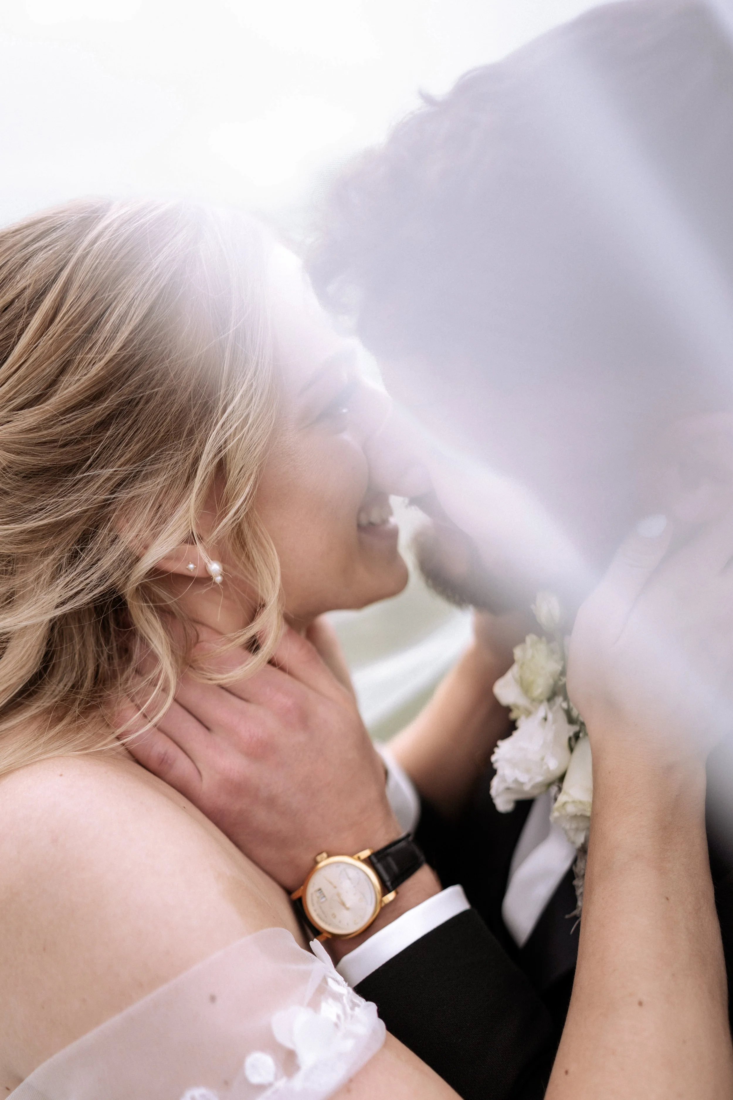 Ein verliebtes Paar bei einer Hochzeitszeremonie, umarmt sich und lächelt, während sie sich nahekommen, mit weißen Blumen und einem weißen Kleid im Hintergrund.