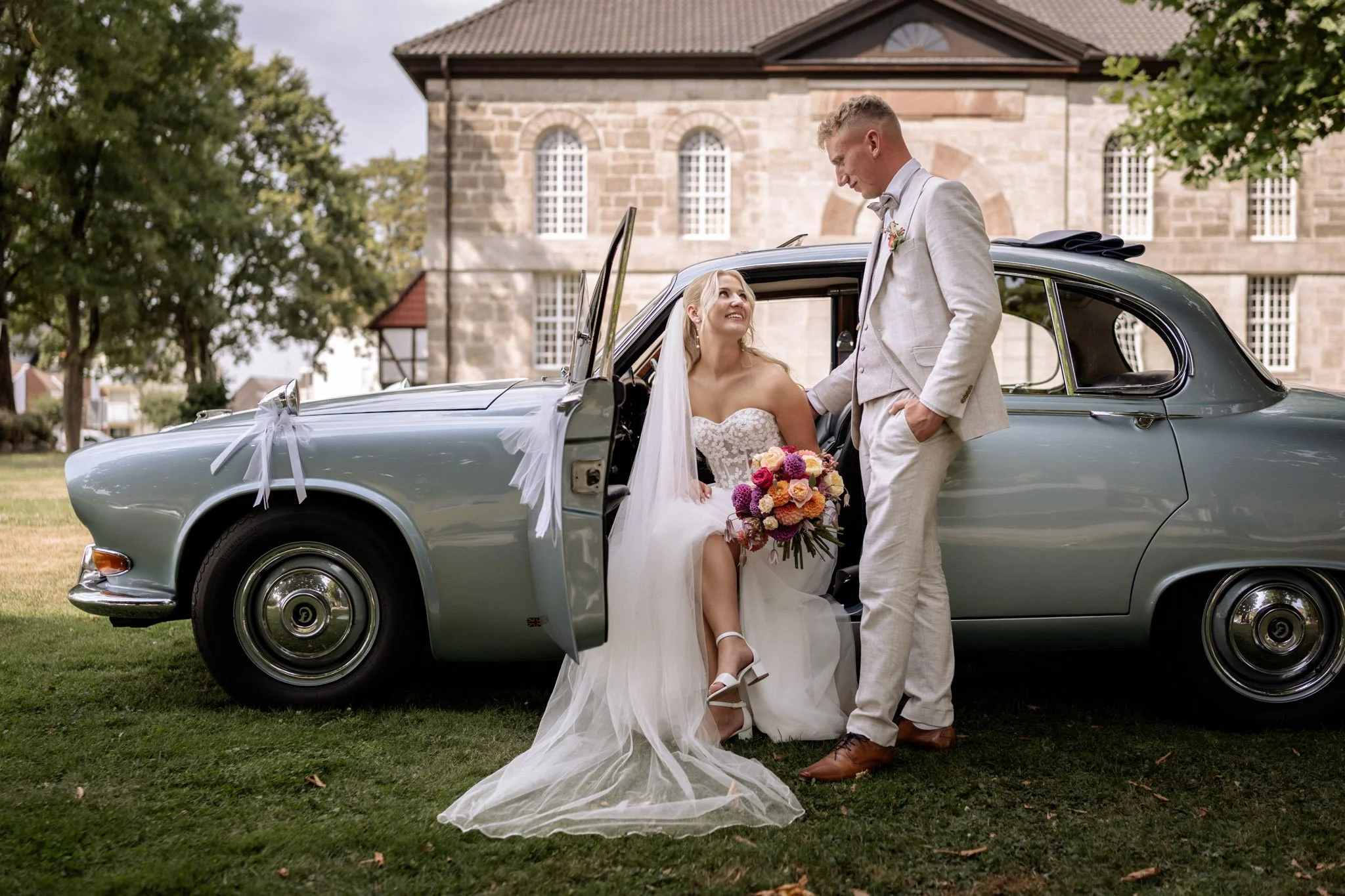 Braut und Bräutigam stehen vor einem klassischen, grauen Auto mit weißen Schleifen, vor einer historischen Backstein-Kirche, im Freien, bei einer Hochzeit.