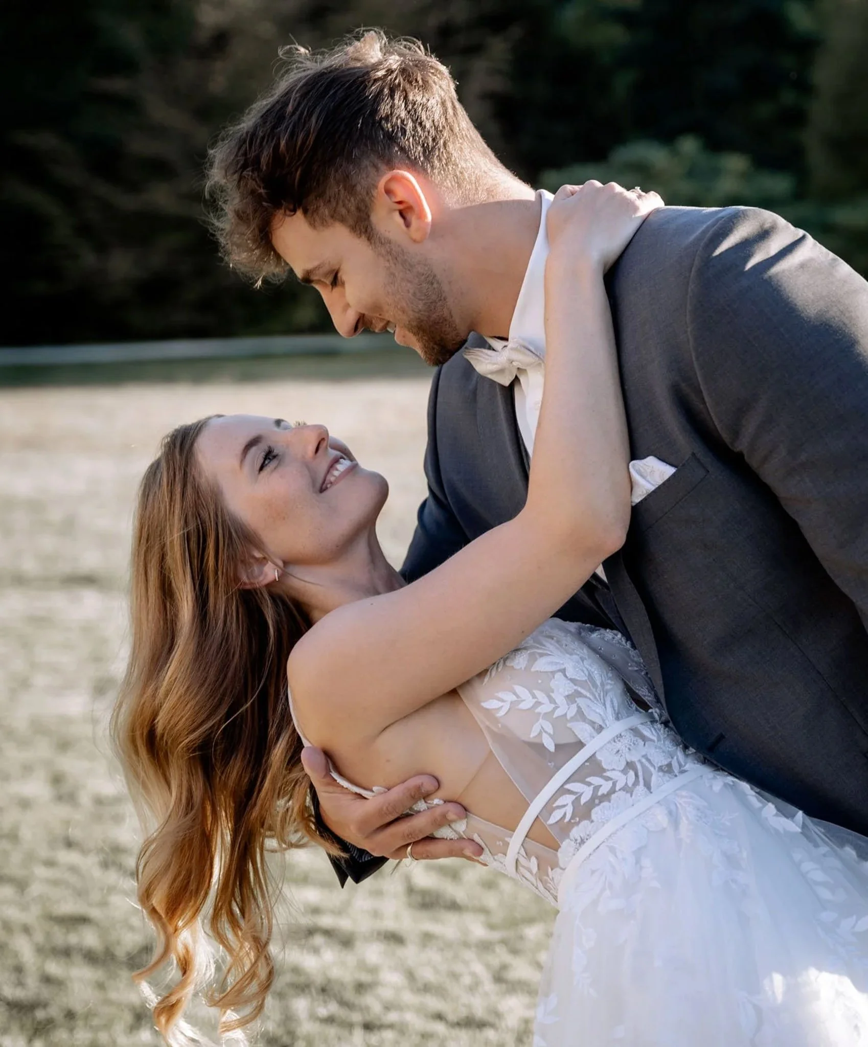 Ein glückliches Paar bei ihrer Hochzeit im Freien, die Frau trägt ein weißes Brautkleid mit Blumenmuster und der Mann einen schwarzen Anzug mit Fliege, sie lachen und schauen sich verliebt an.