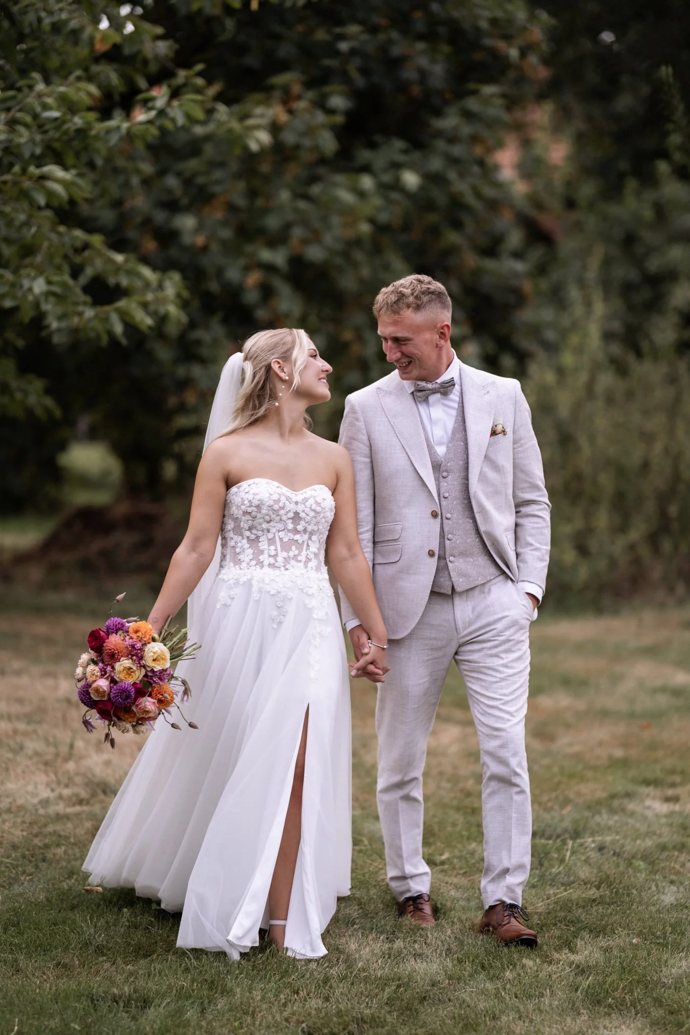 Braut und Bräutigam beim Spazieren im Grünen, lächelnd und Hand haltend. Die Braut trägt ein weißes Hochzeitskleid mit Blumenmuster, die Brautjungfer hält einen bunten Blumenstrauß.