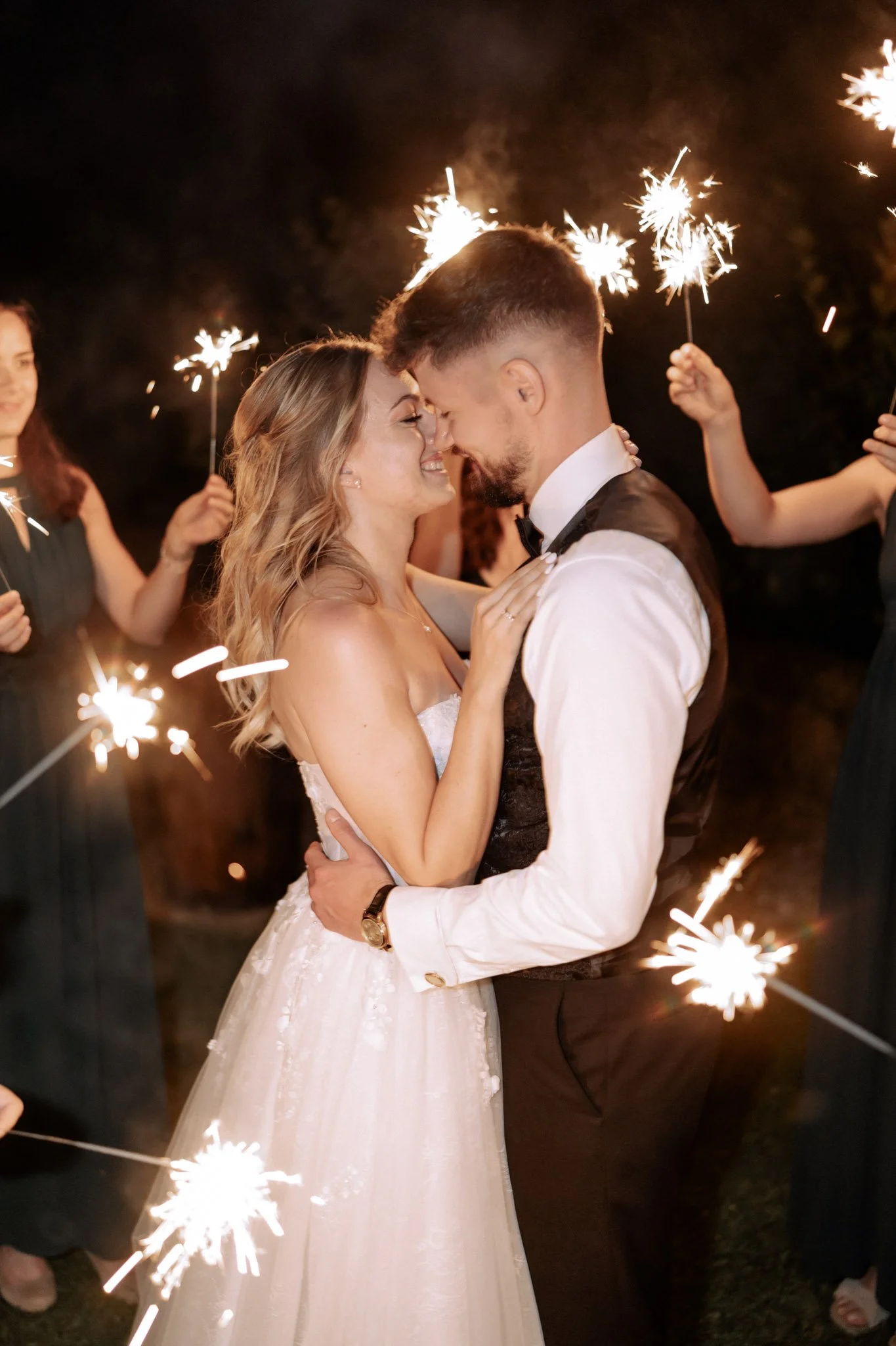 Ein Paar, das bei einer Hochzeit zärtlich zusammen tanzt, umgeben von Freunden, die mit Feuerwerkskörpern feiern.