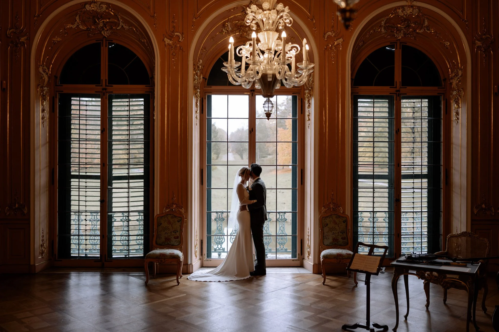 Ein Brautpaar, das sich liebevoll umarmt, steht in einem eleganten, opulent verzierten Raum vor großen Fenstern, durch die ein herbstlicher Garten sichtbar ist, mit einem prunkvollen Kronleuchter hangs daran.