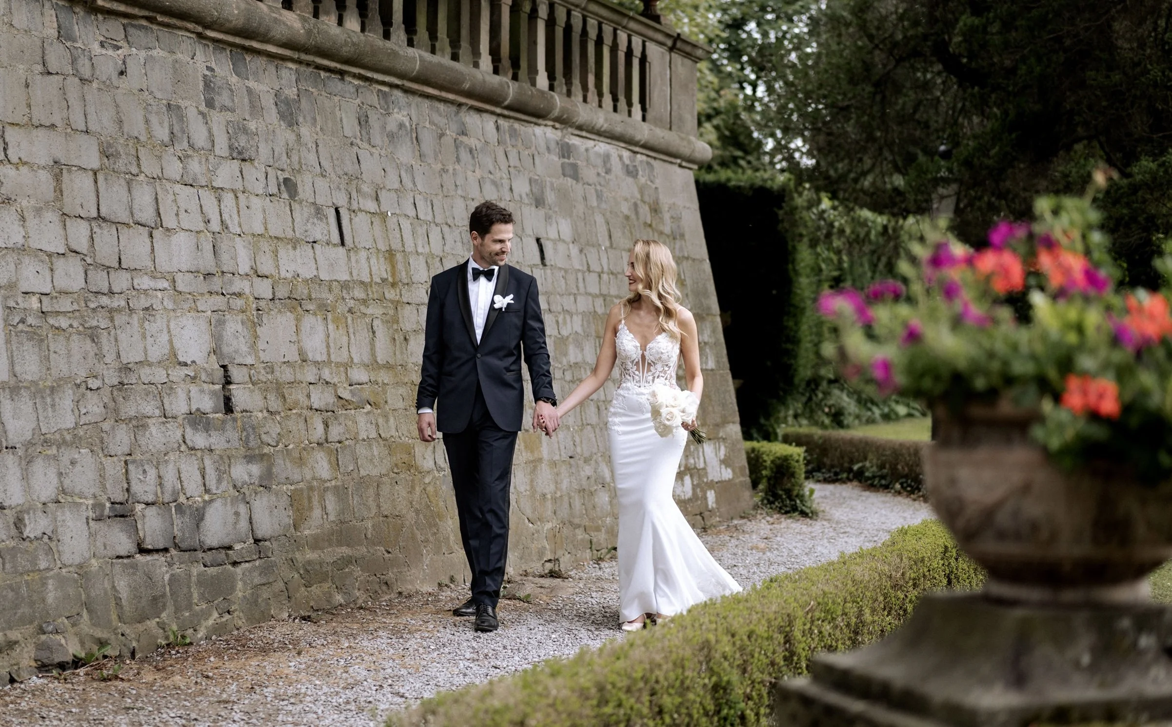 Ein Brautpaar in formeller Kleidung, die Hand in Hand spazieren gehen, im Garten mit Blumen und einer Steinmauer im Hintergrund.